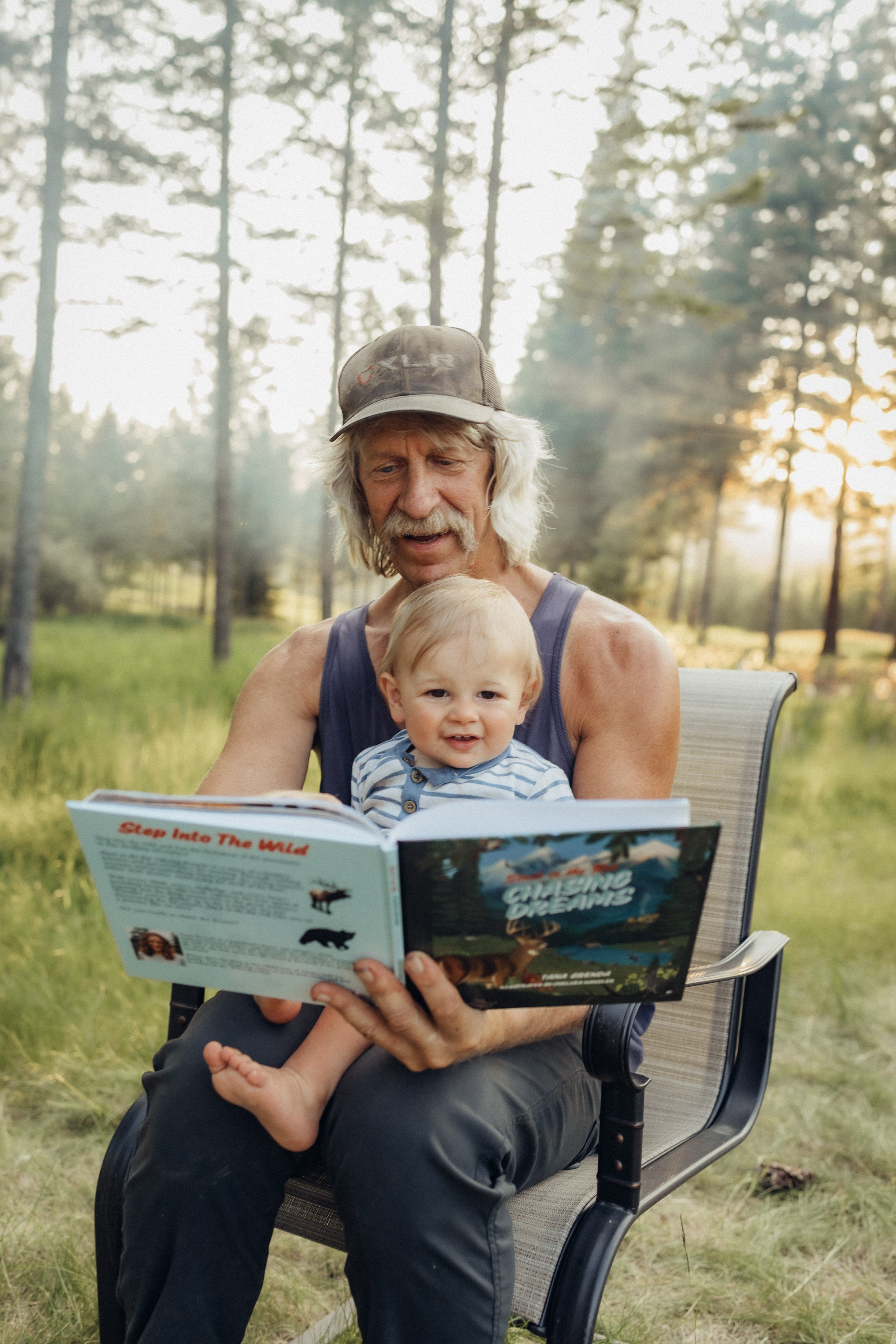 An elderly man with long gray hair and a mustache reading a book titled "Guiding Dreams" to a toddler sitting on his lap outdoors in a forested area during sunset.