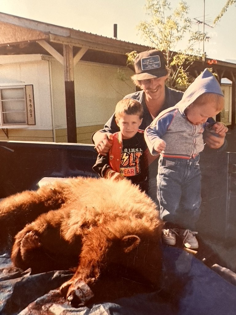 A man with two young children standing next to a lion that appears to be dead, lying on its side, on the bed of a truck. The man is smiling, one child is wearing a gray hoodie, and the other child is holding an orange strap. The setting appears to be