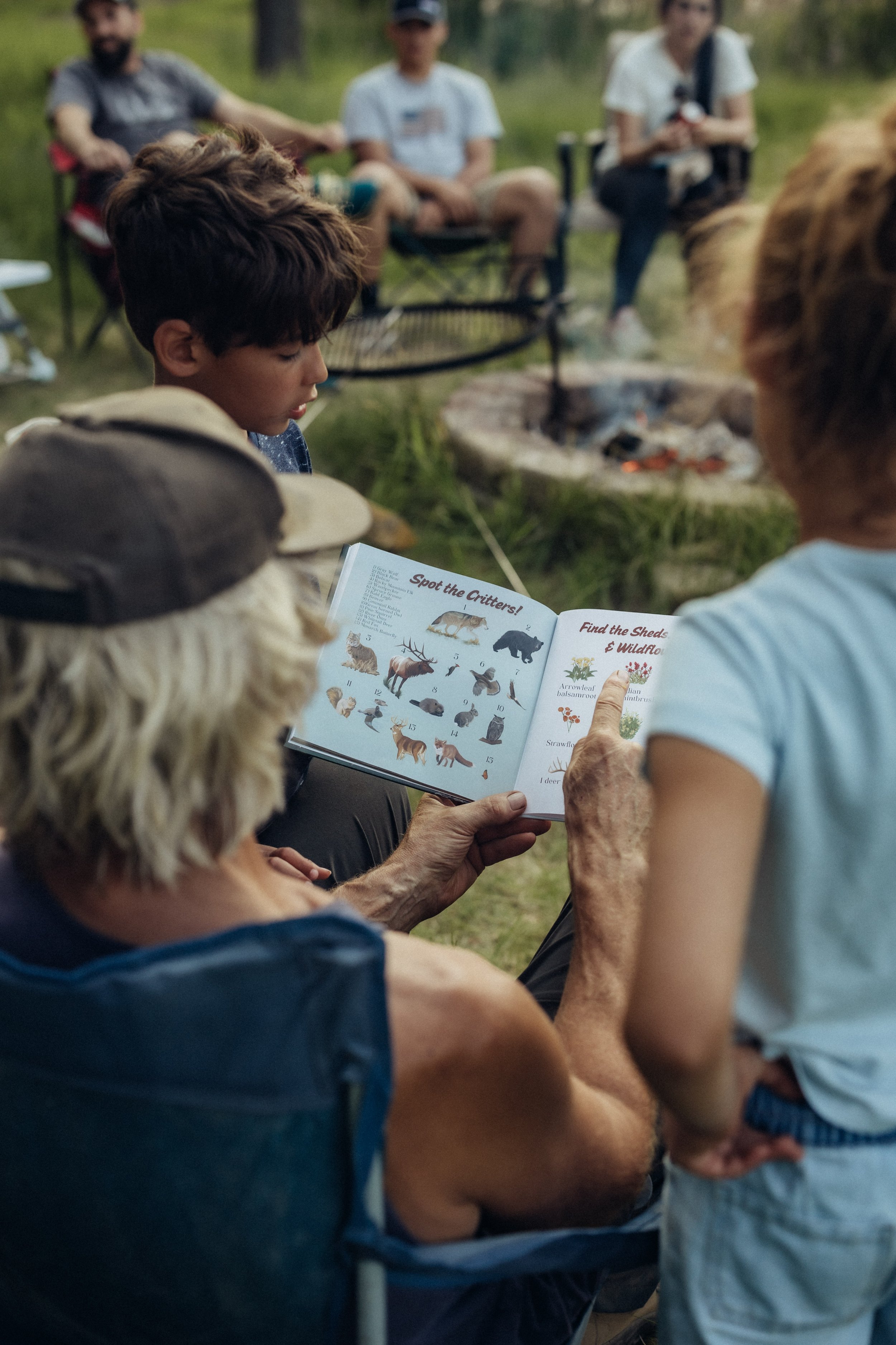 Older woman with gray hair reading a children's book about wildlife to children sitting around a campfire outdoors at dusk.