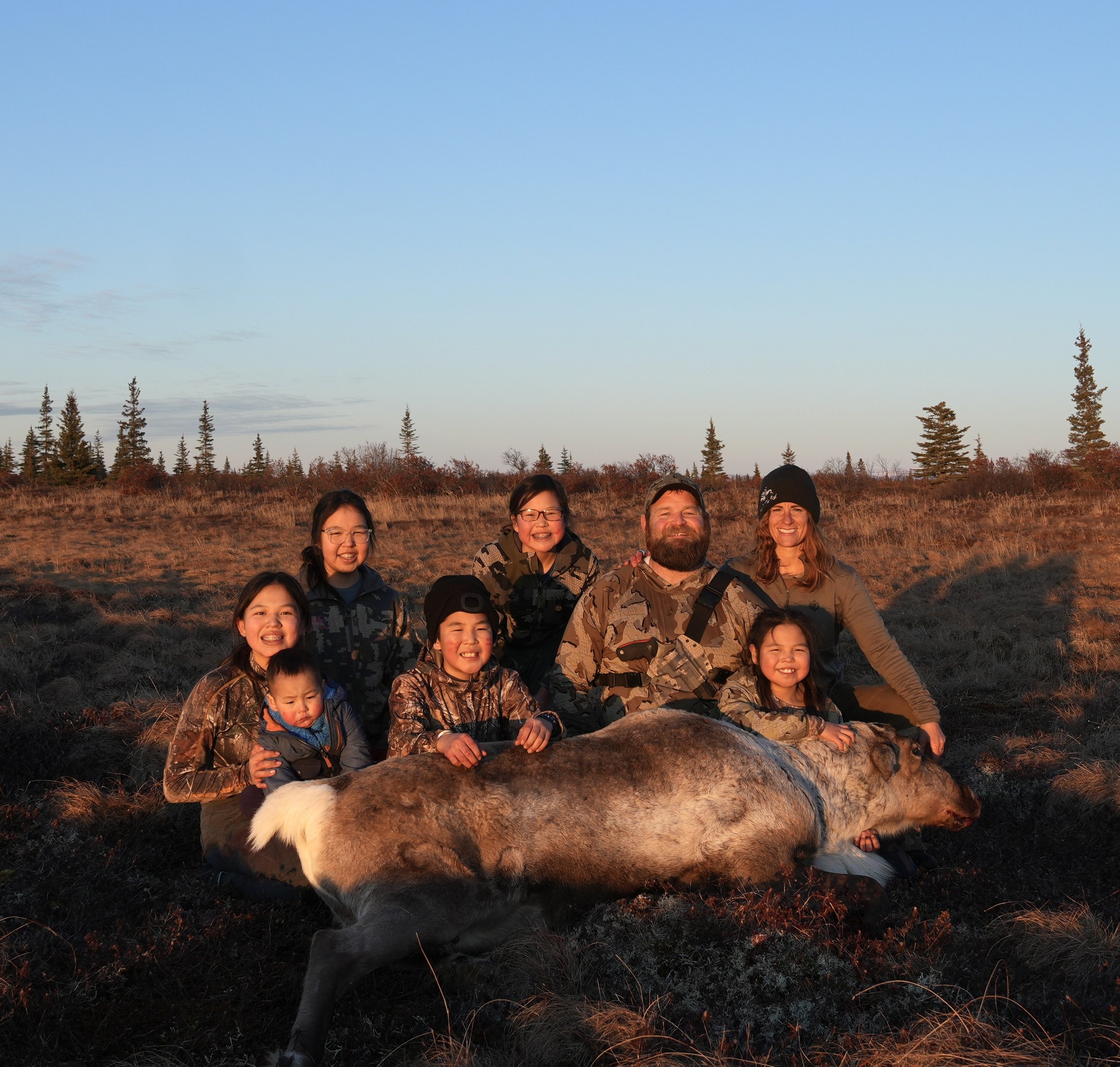 Group of seven people posing with a dead elk in a field during sunset, wearing camouflage clothing and outdoor gear.