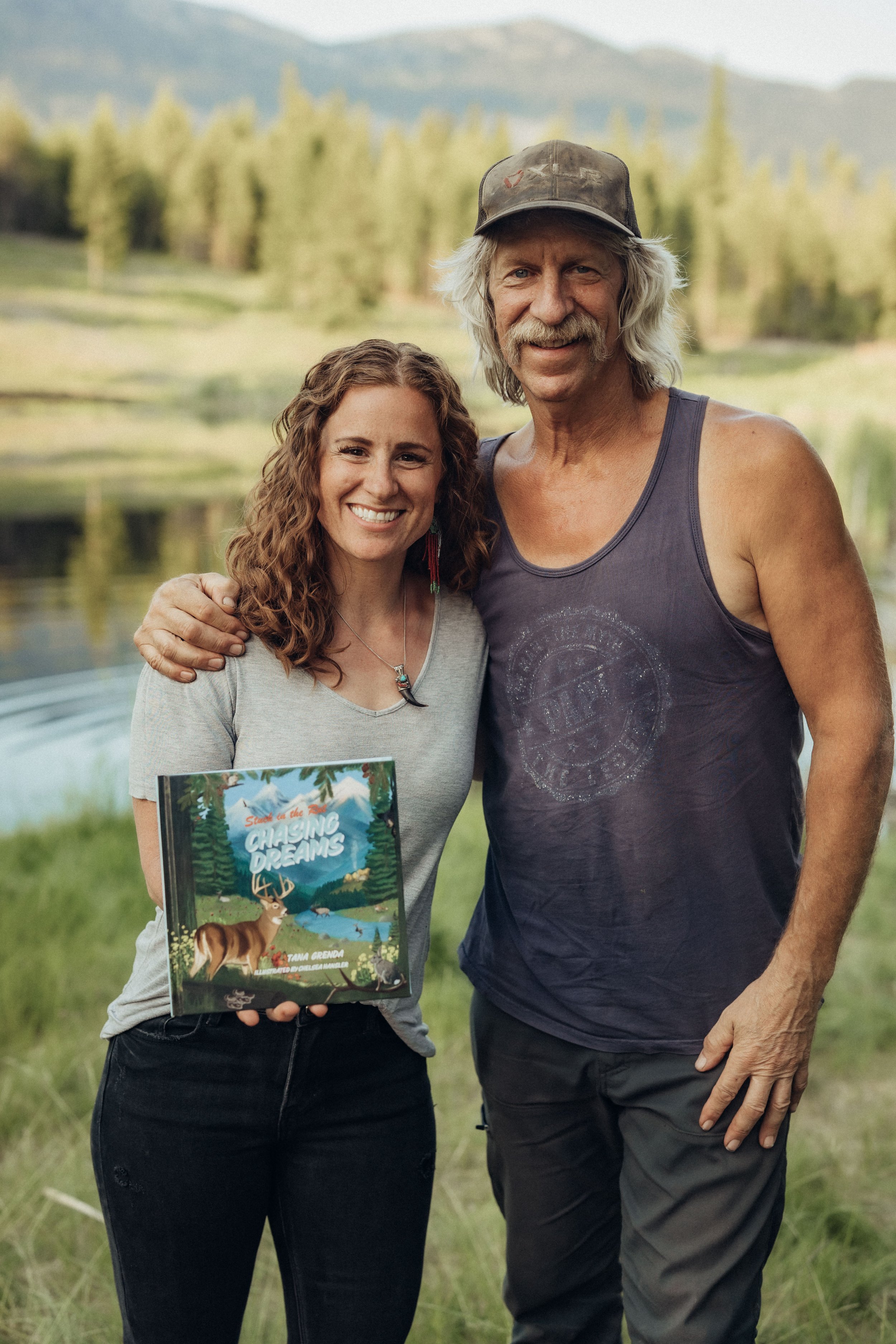 A woman and a man standing outdoors by a lake, both smiling, with the woman holding a children's book titled "Catching Dreams" featuring a deer on the cover.