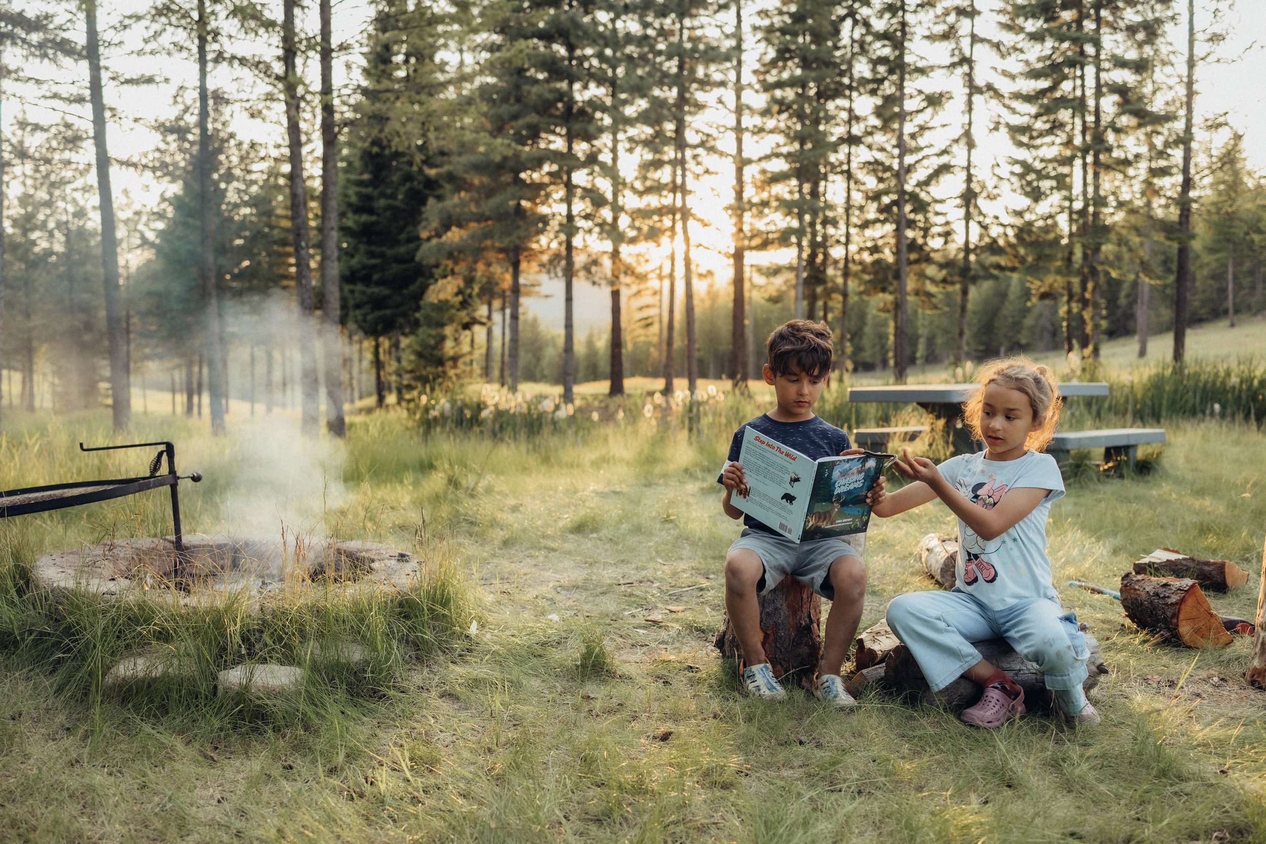 Two children sitting on logs in a forest clearing, reading a book together during sunset.