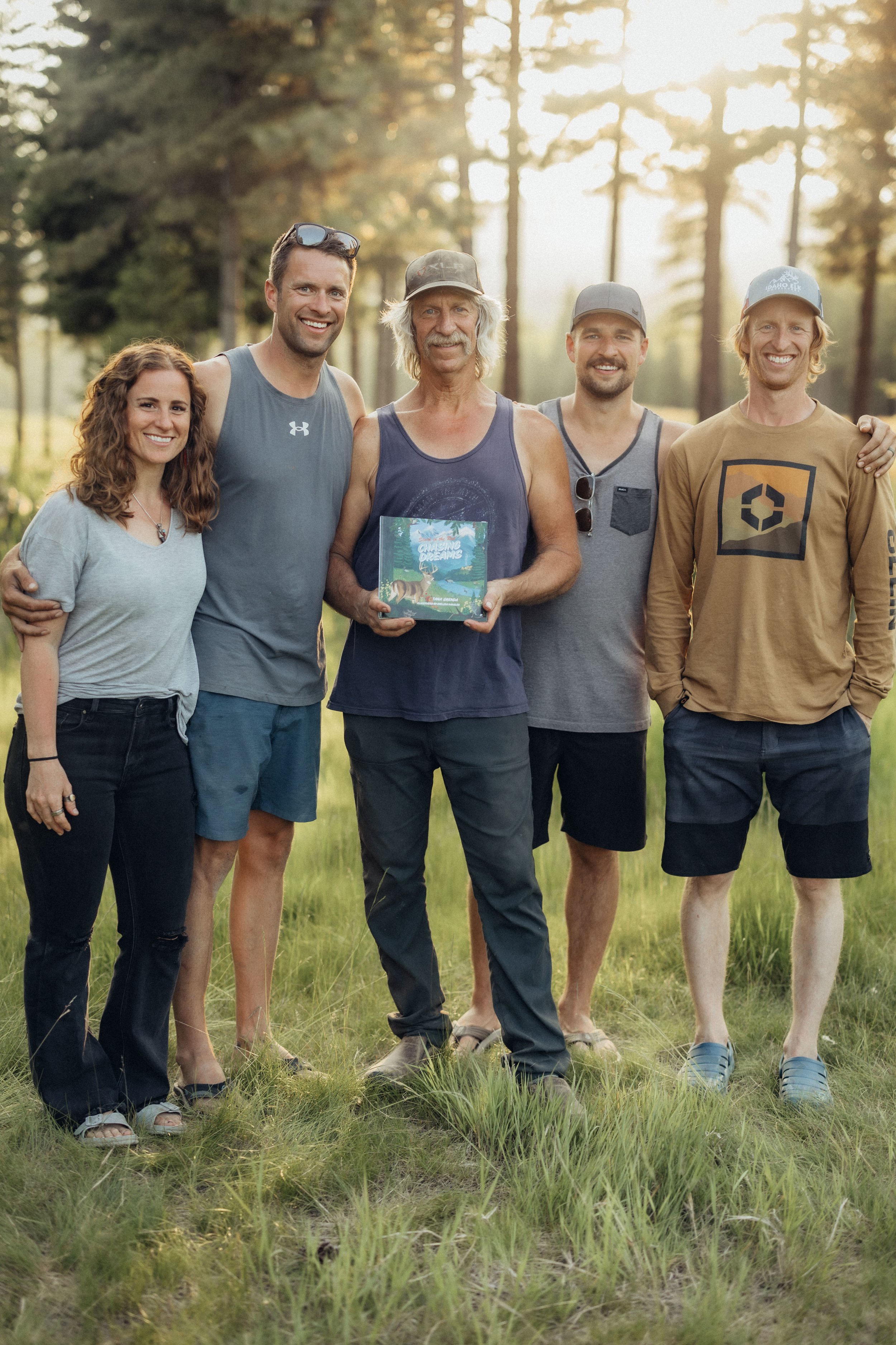 A group of five friends standing outdoors in a grassy area with trees and sunlight in the background, smiling and posing for the photo.