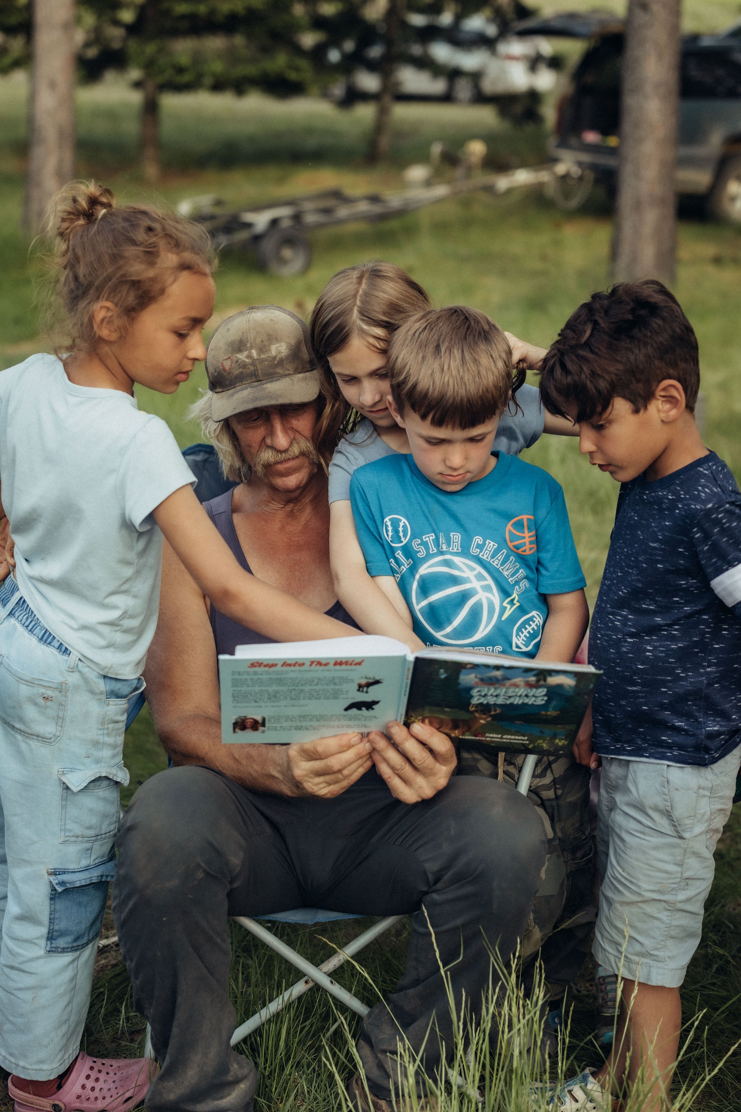 An elderly man with a gray beard and cap reading a book titled 'Stop Into The Wild' to four children gathered around him outdoors in a grassy area with trees and a vehicle in the background.