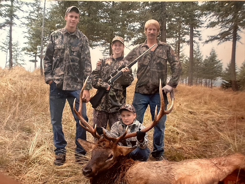 A family of four dressed in camouflage in a forest, posing with a large buck they hunted. The group includes a woman, two boys, and a man, all smiling and standing behind the deer.