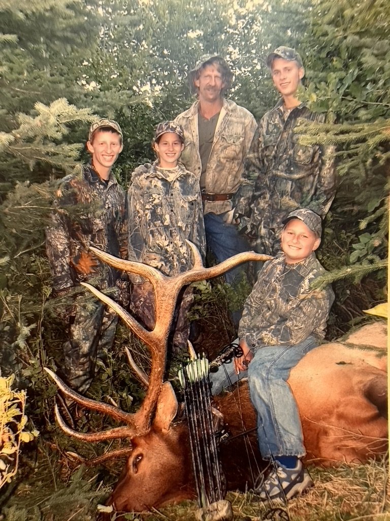 A family of five dressed in camouflage hunting gear posing outdoors behind a large antlered deer they have hunted, with a bow resting on the deer.