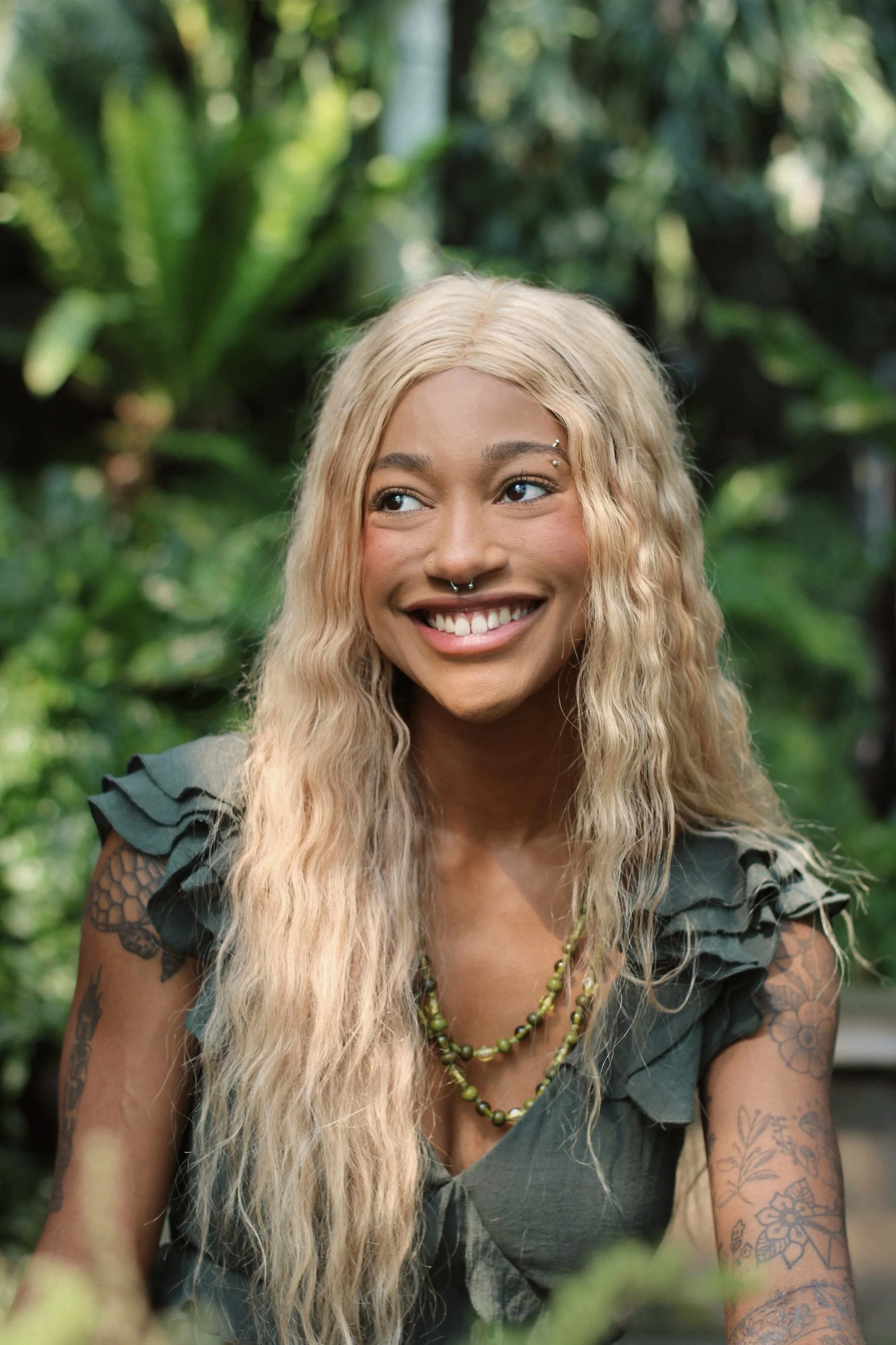 A smiling woman with long, wavy blonde hair and tattoos on her arms, outdoors with lush green foliage in the background.