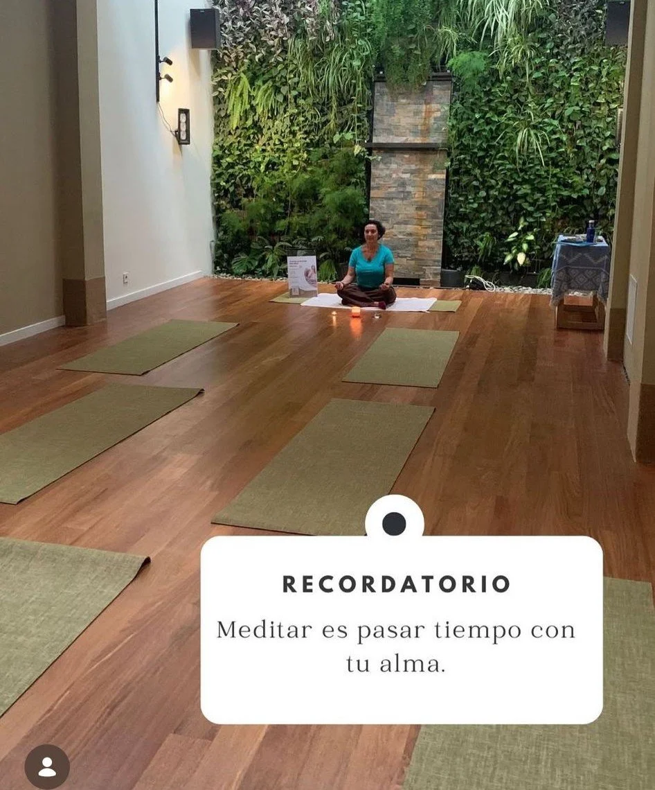 Una mujer sentada en posición de meditación en un salón con paredes verdes, piso de madera y un ambiente tranquilo.