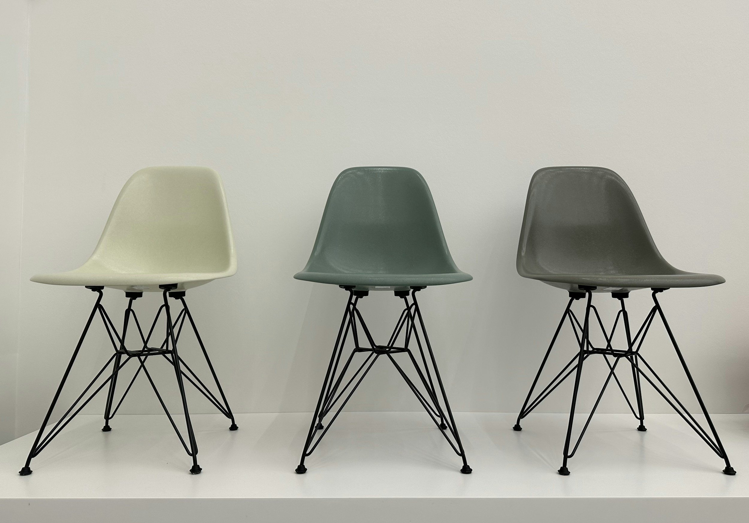 Three Eames modern chairs with black wire legs in beige, green, and gray, arranged in a row on a white surface against a white wall.