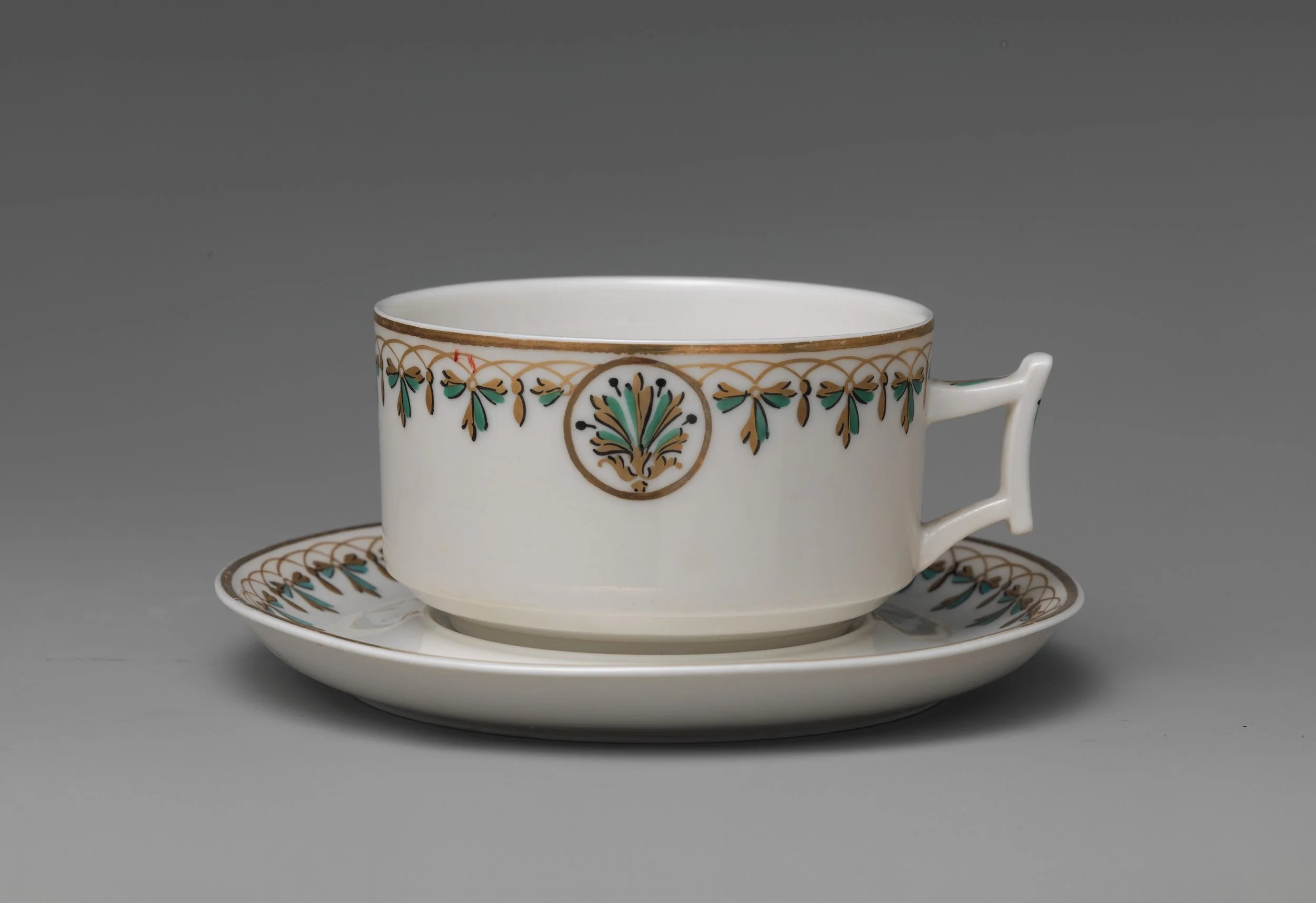 Porcelain teacup with ornate gold and green decoration, placed on matching saucer and clear glass plate, against a plain gray background.