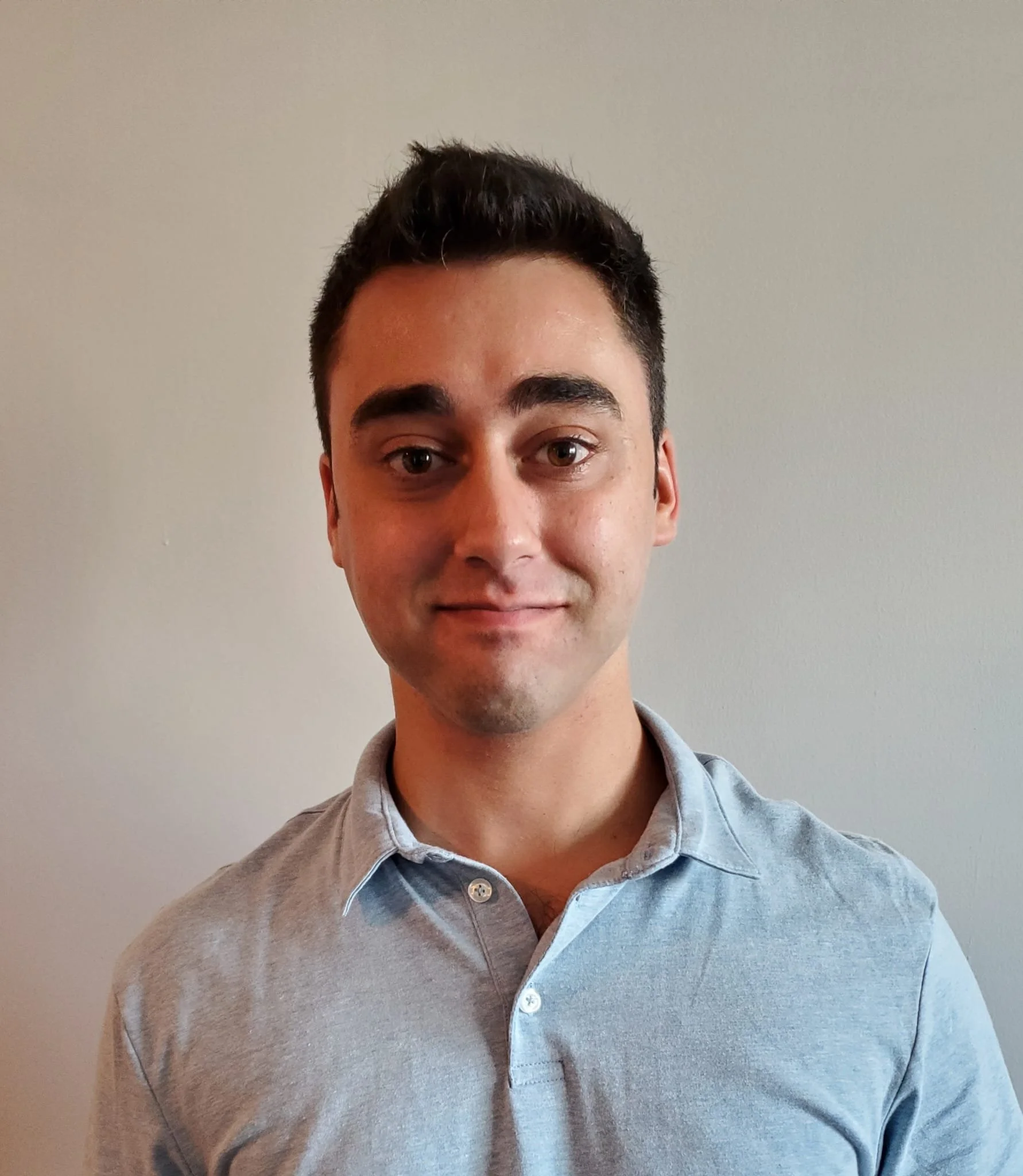 A young man with short dark hair and fair skin, wearing a light grey collared shirt, standing against a plain off-white wall and smiling gently at the camera.