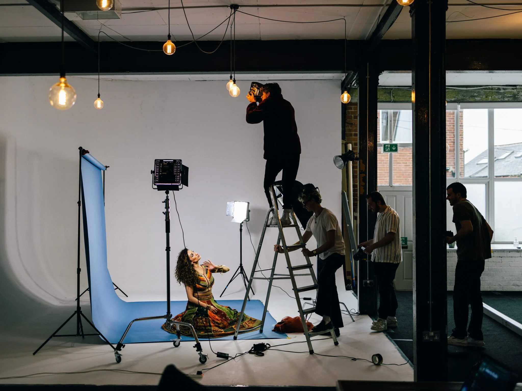 Photography studio with a model on a blue backdrop, a photographer on a ladder taking pictures, and assistants adjusting equipment under hanging lights.
