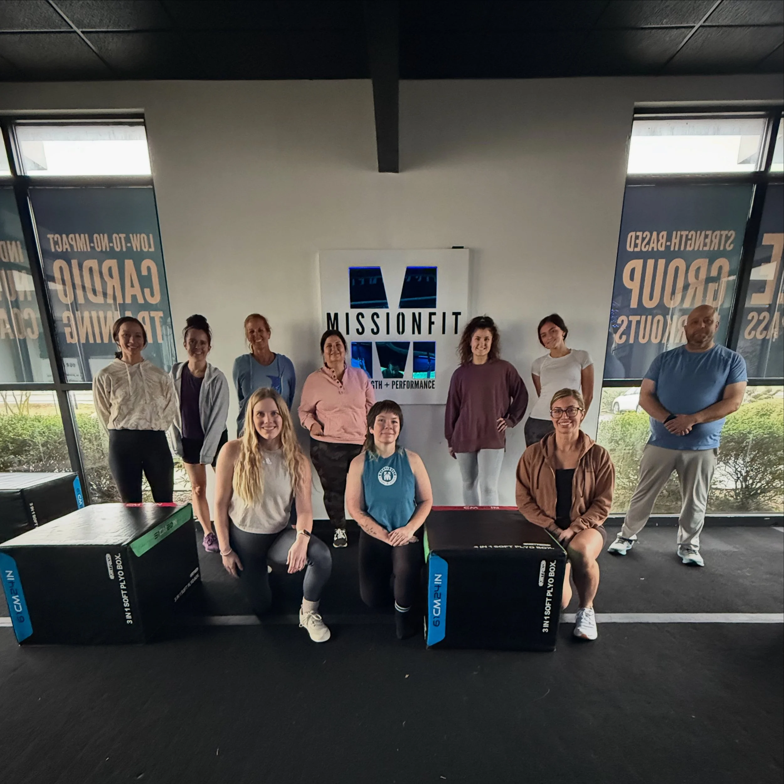 Group of ten people standing in front of MissionFit sign inside gym, with large windows and banners promoting strength-based training.