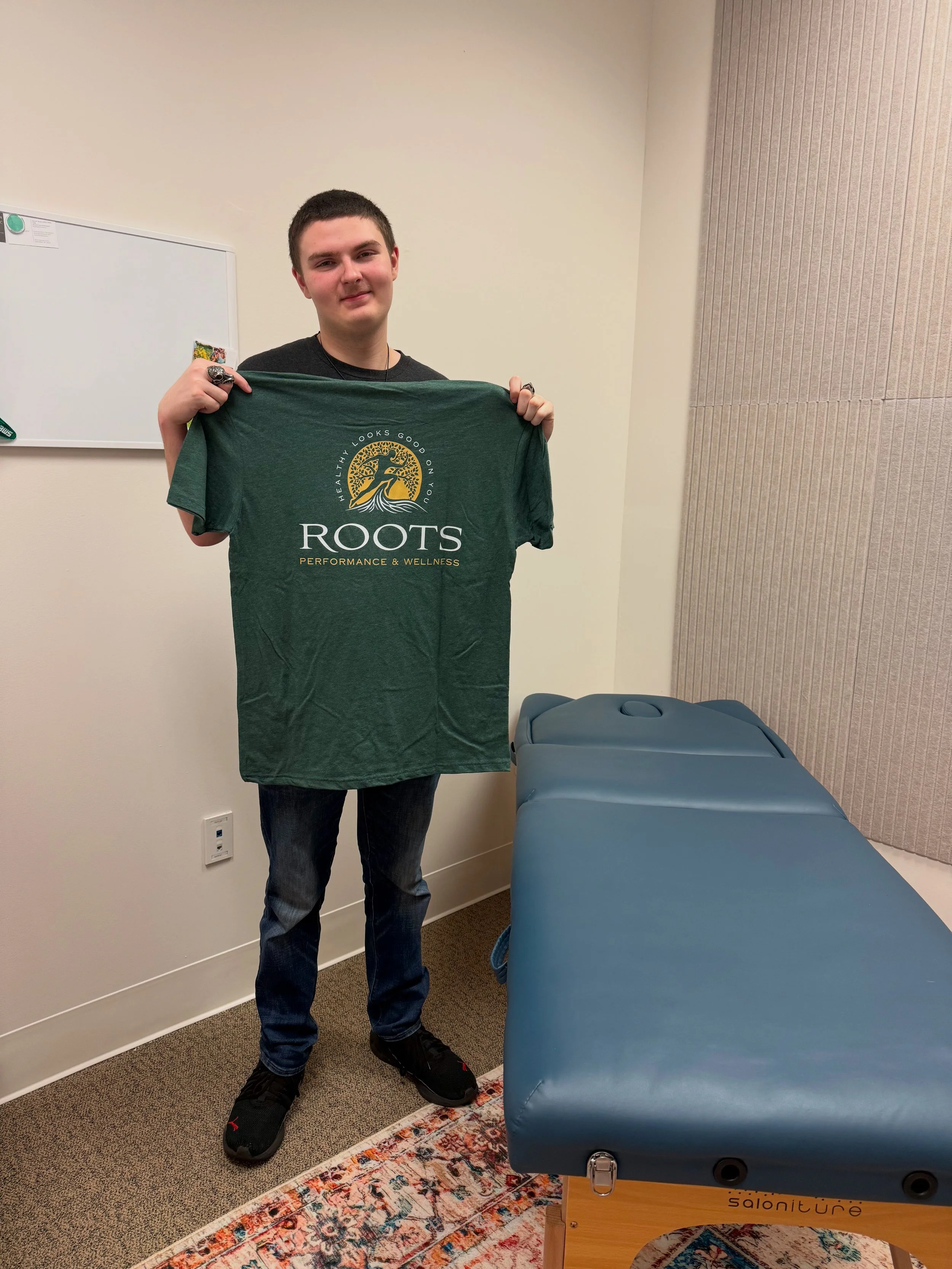 A young man with short dark hair wearing a black shirt, jeans, and black shoes standing in a room, holding a green T-shirt with a logo and text that reads 'ROOTS PERFORMANCE & WELLNESS.' The room has beige walls in physical therapy clinic.