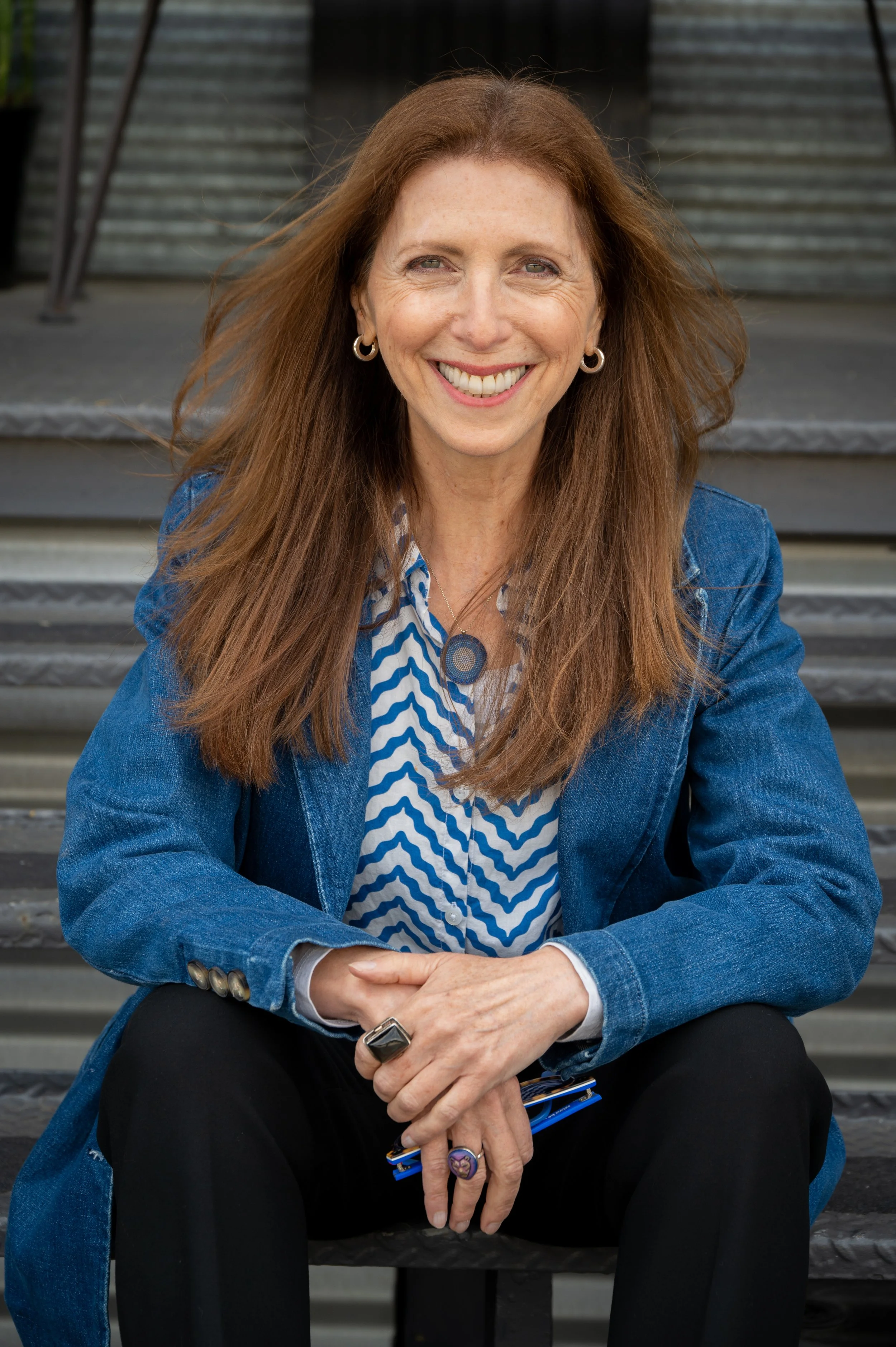 A woman with long red hair smiling, wearing a blue denim jacket and a patterned shirt, sitting on stairs.