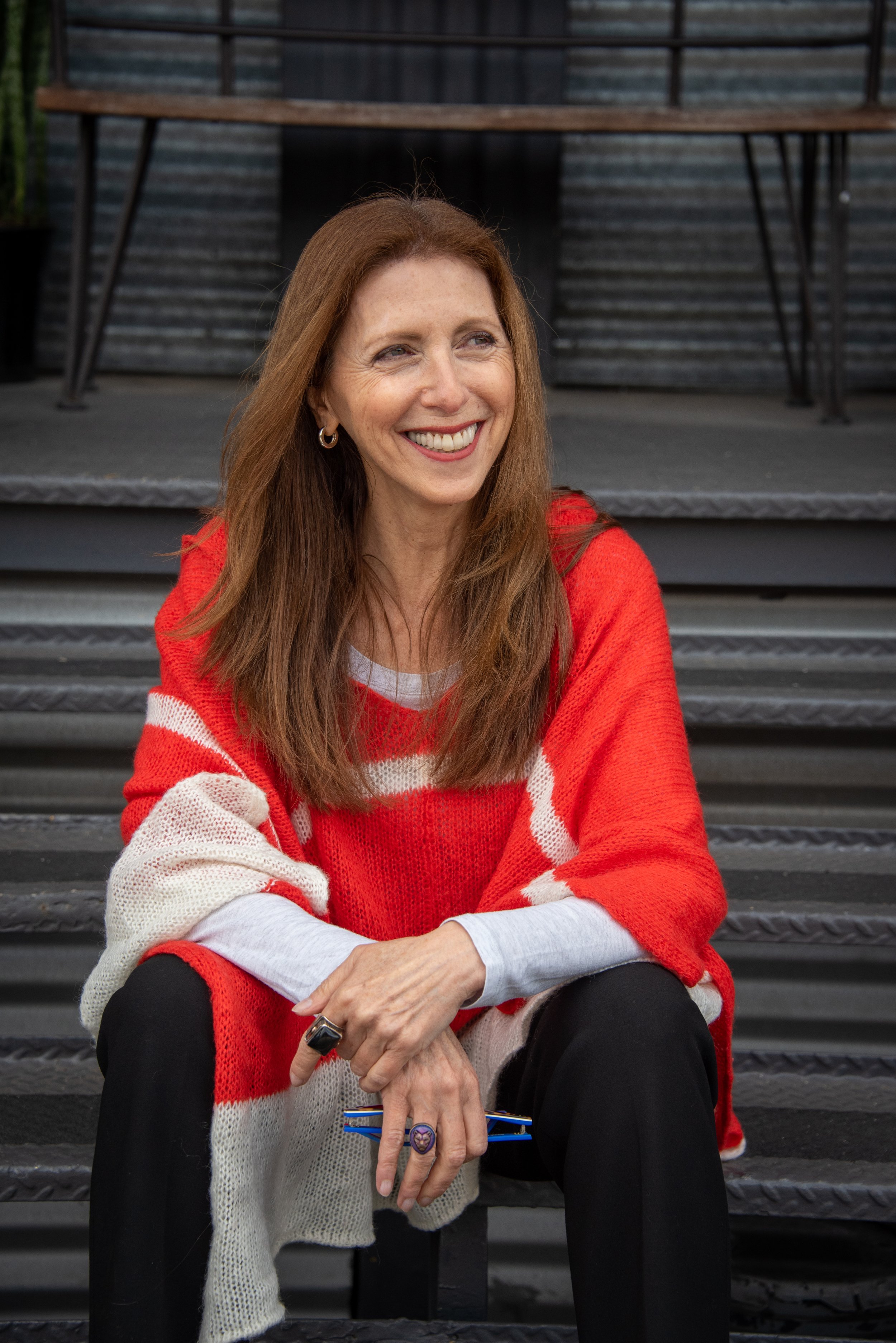 A smiling woman with long red hair, wearing a red and white sweater, sitting on outdoor stairs.