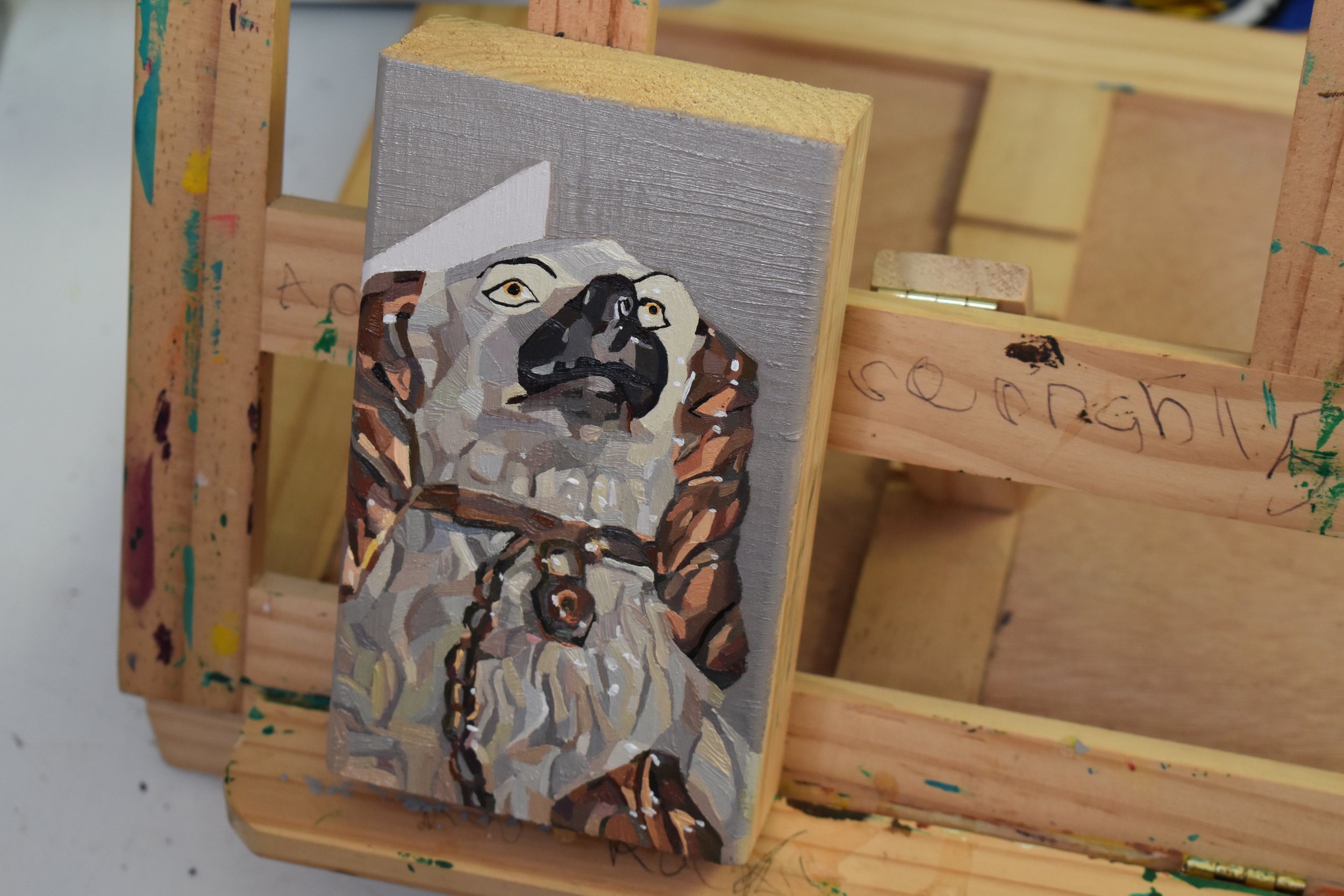 Painted wooden block featuring a stylized picture of a ceramic dog ornament, set against a grey background.