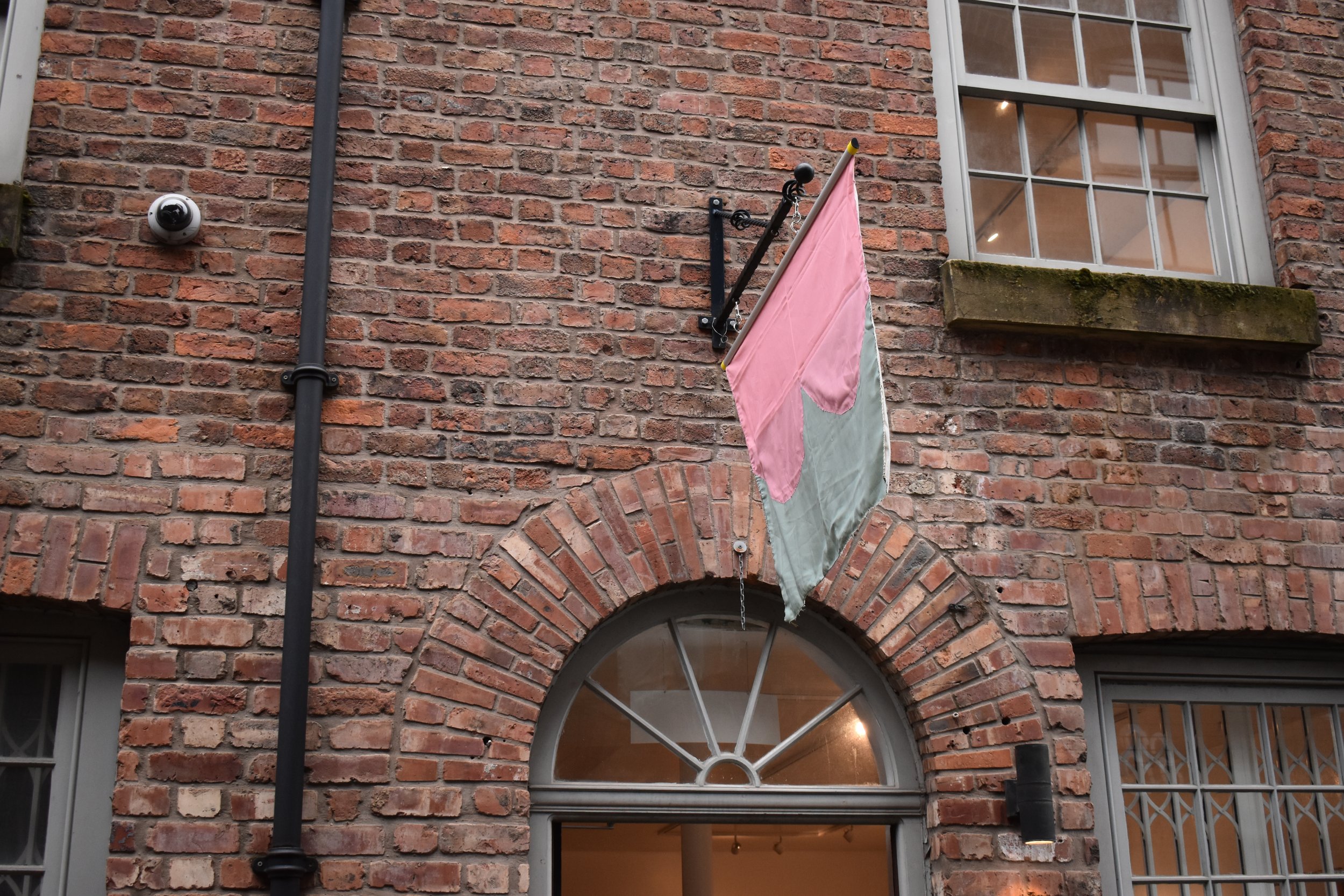 A brick building with a pink and gray fabric flag hanging from a metal pole above a rounded window door.