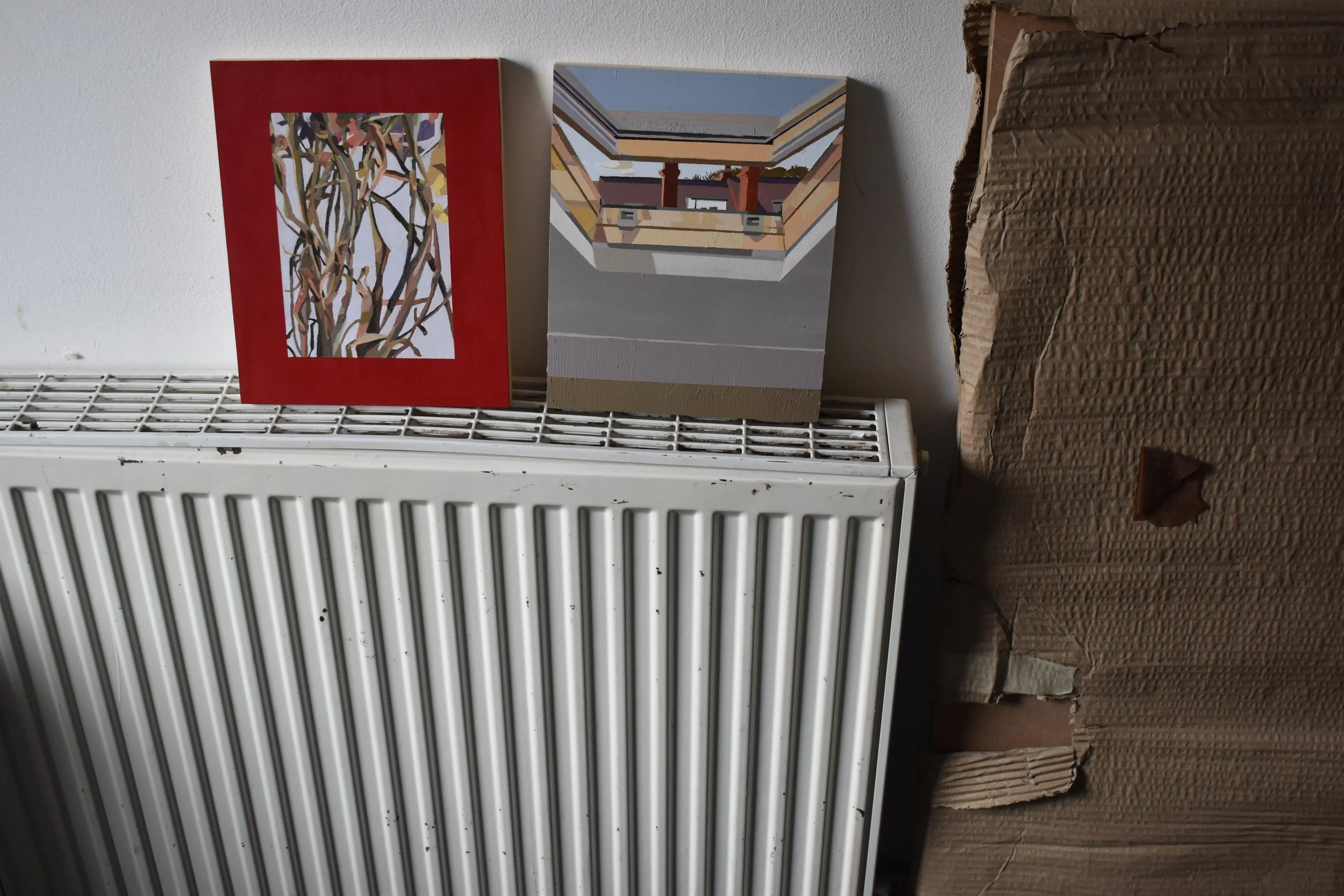 Two small paintings rest on a white radiator; one features abstract branches with pink and purple leaves surrounded by a red border, the other depicts a geometric perspective of a building with a blue sky in the background.