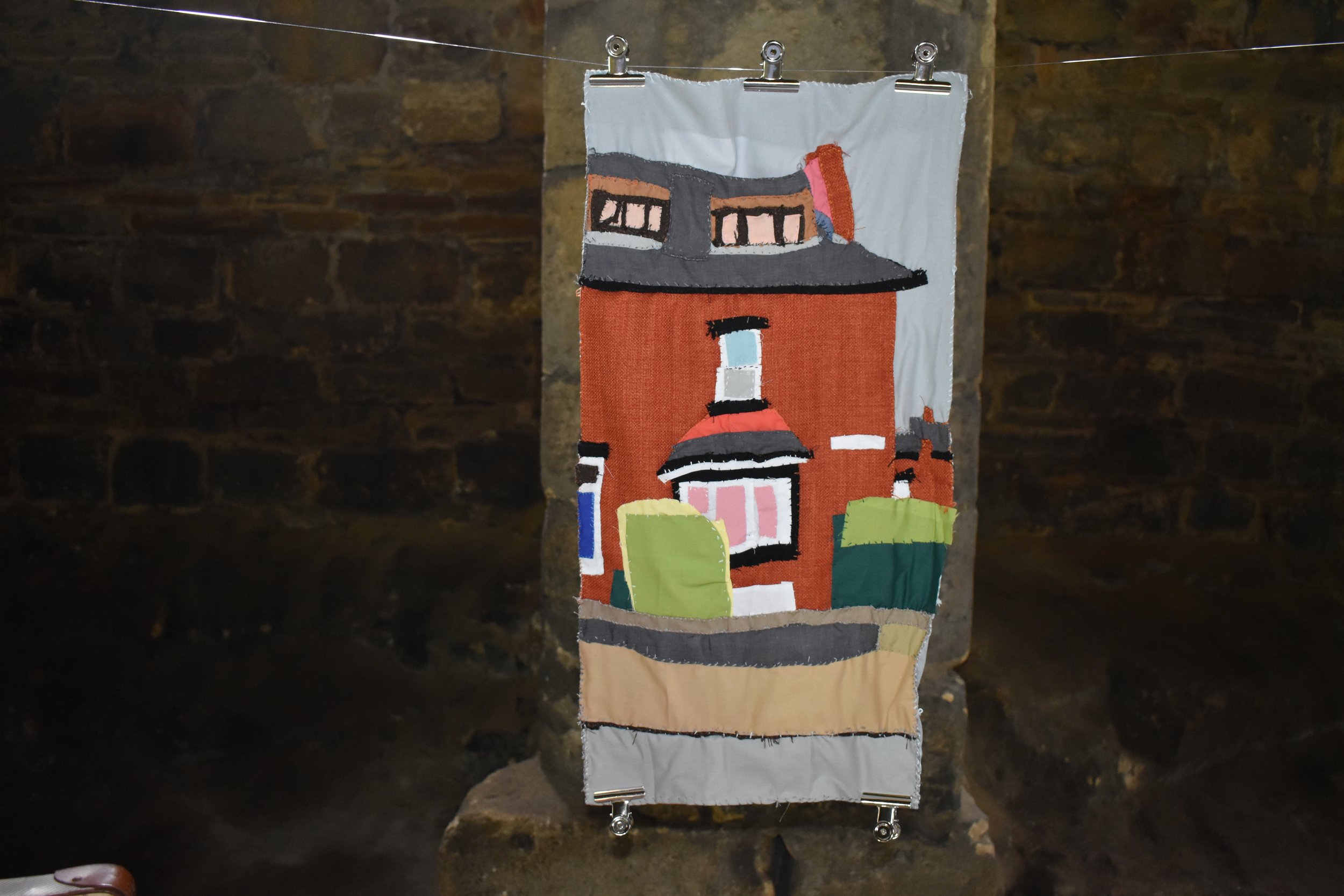 A textile wall hanging depicting a house with a red brick exterior, black roof, and windows, attached to a string with clips, set against a rustic stone wall background.