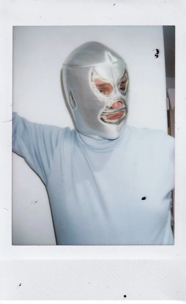 Person wearing a silver luchador mask and a light-colored t-shirt, standing against a white background.
