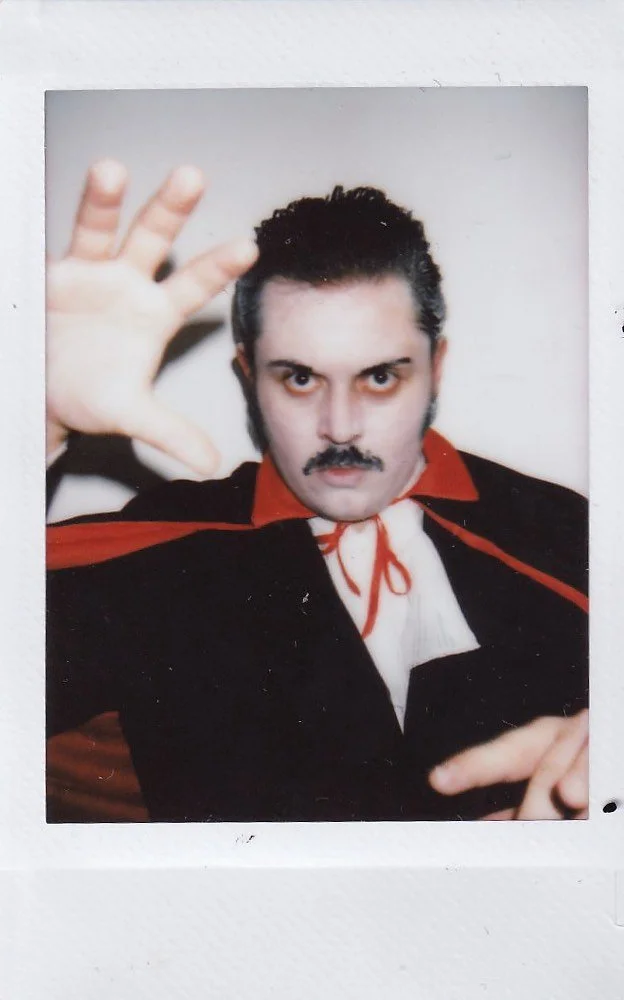 A person dressed as a vampire with a cape, white shirt, and red bow tie, reaching towards the camera with one hand, and pointing with the other, with a serious facial expression and dark hair and mustache.