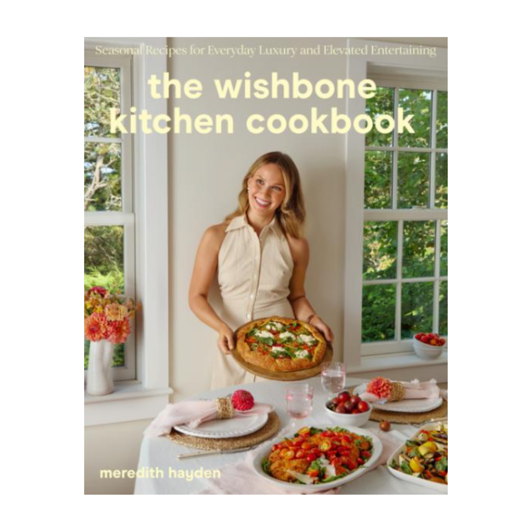 Cover of the Wishbone Kitchen Cookbook by Meredith Hayden featuring a woman holding a pizza in a bright kitchen with windows and a table set with salads and dishes.
