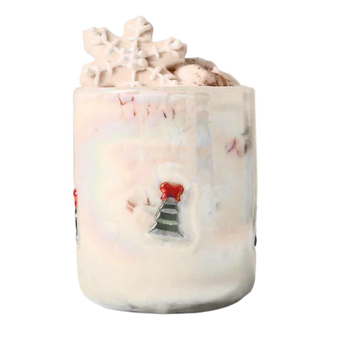 A festive clear glass filled with a creamy white holiday beverage, topped with whipped cream, a snowflake-shaped cookie, and sprinkles, with holiday-themed decorations on the glass.