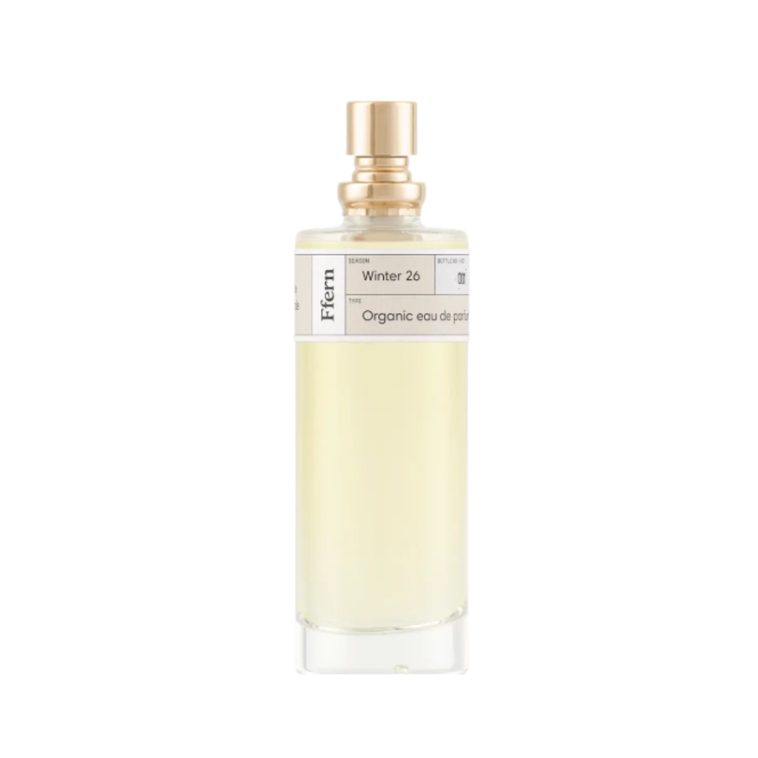 A clear glass bottle with a gold cap containing a pale yellow liquid, labeled with a white tag reading 'Winter 26,' 'Organic eau de parfum,' and 'Fern.'