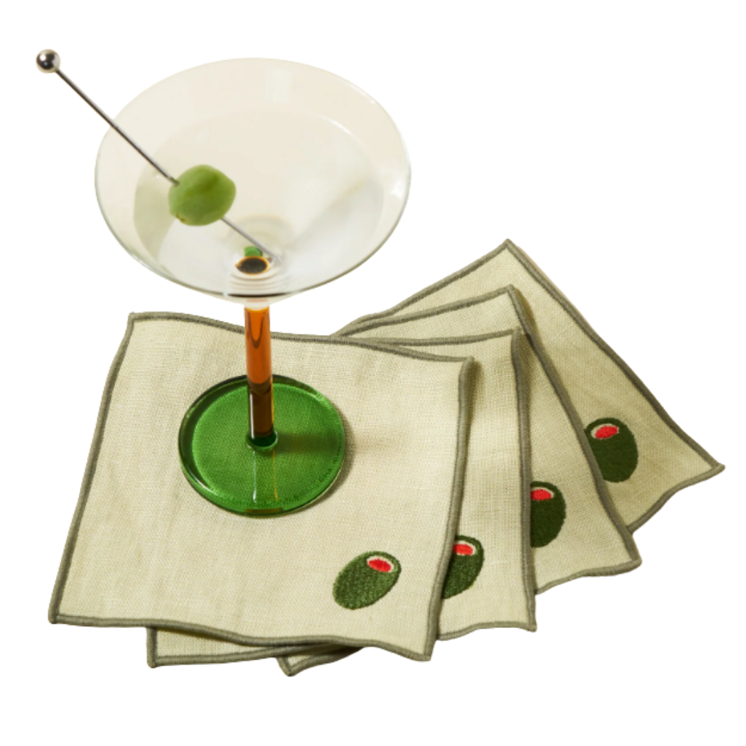 A clear martini glass with an olive on a cocktail pick, placed on a beige napkin with olive print, isolated on a white background.