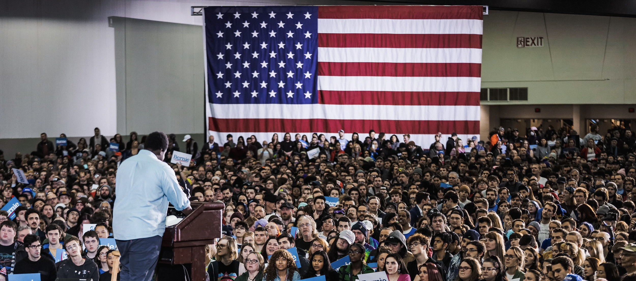 A large crowd of people gathered indoors at a political rally or event, with a prominent large American flag hanging on the wall behind them. A man is speaking at a podium in the foreground, with some attendees holding signs that say "Bernie."