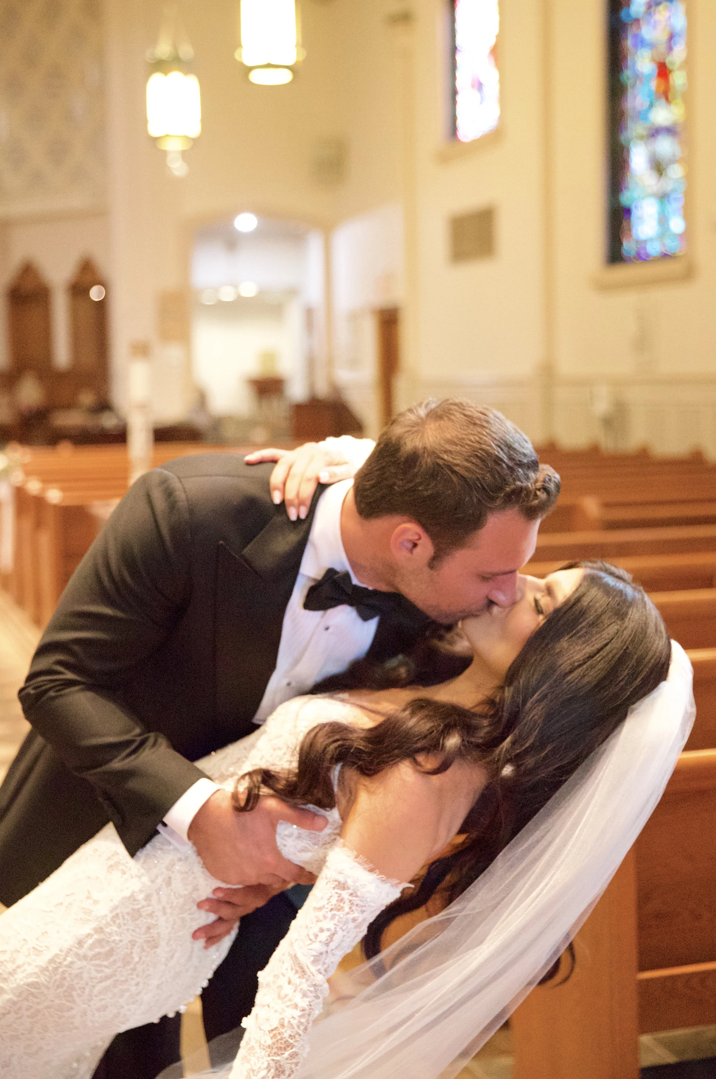 A newlywed couple sharing a kiss inside a church, with the groom in a tuxedo and the bride in a lace wedding dress and veil.