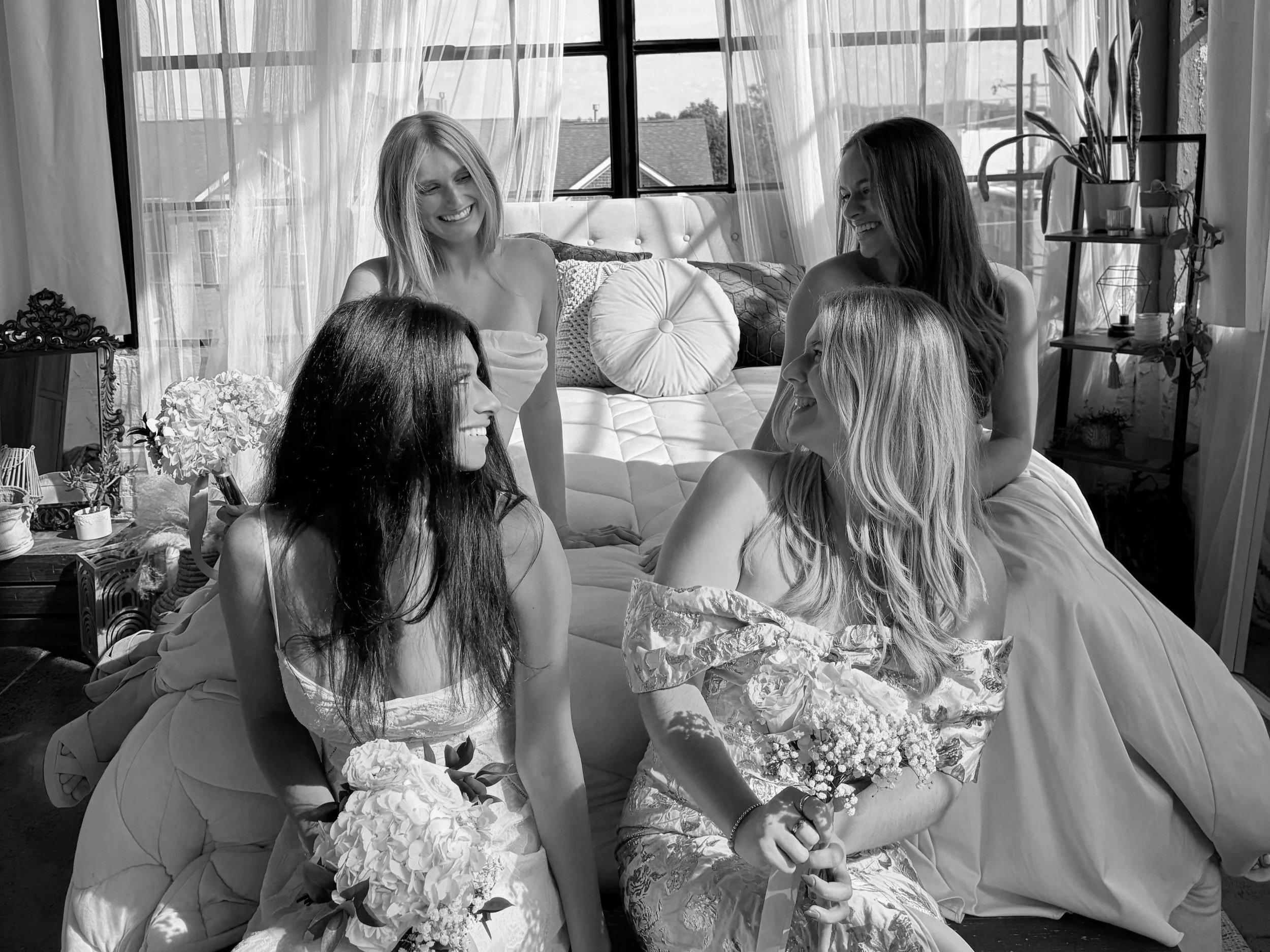 Five women in dresses sitting and standing on a sofa and the floor, smiling and engaging with each other in a sunny room with large windows and potted plants.