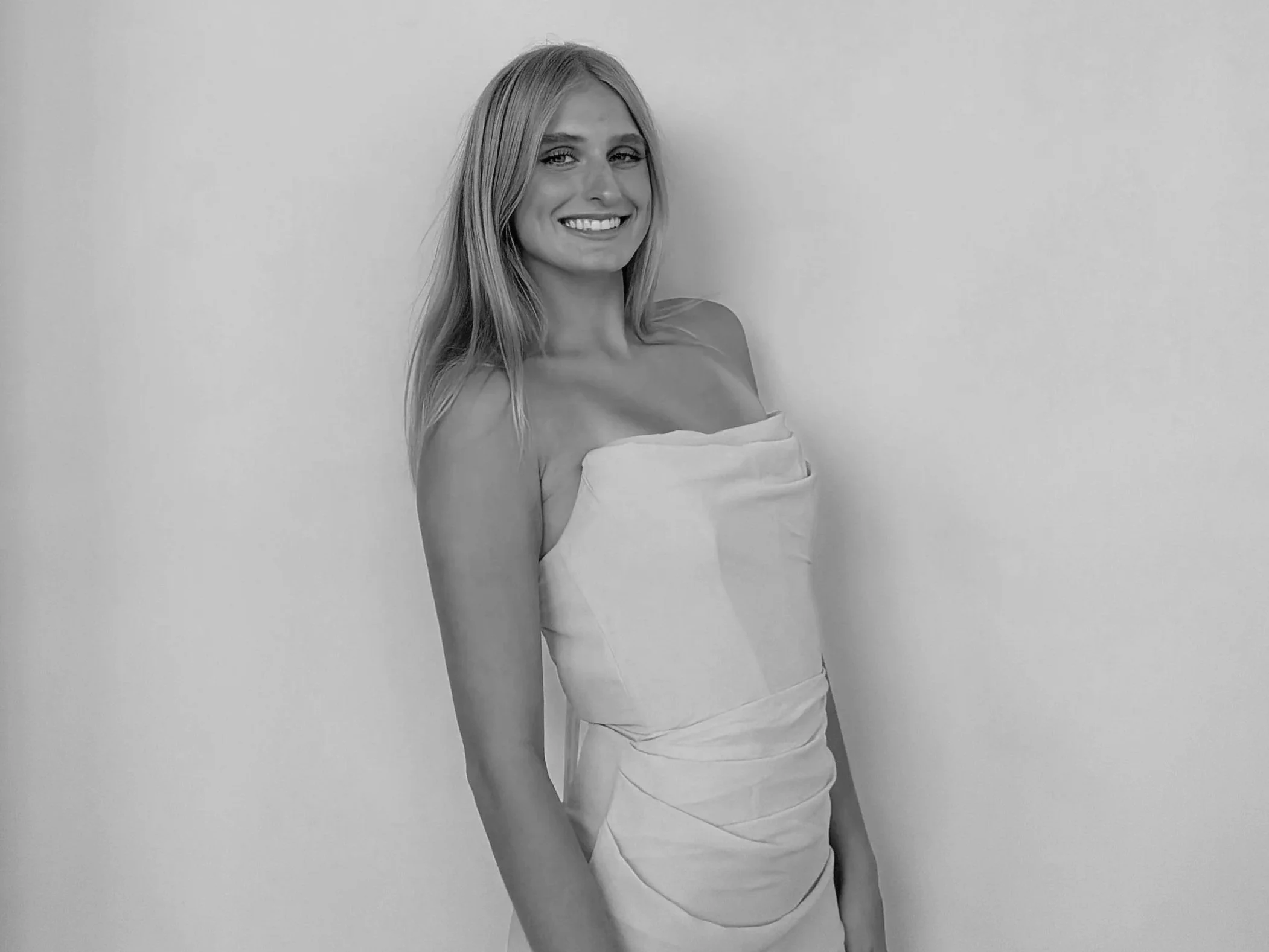 Black and white photo of a smiling young woman with long hair, wearing a strapless dress, standing against a plain wall.