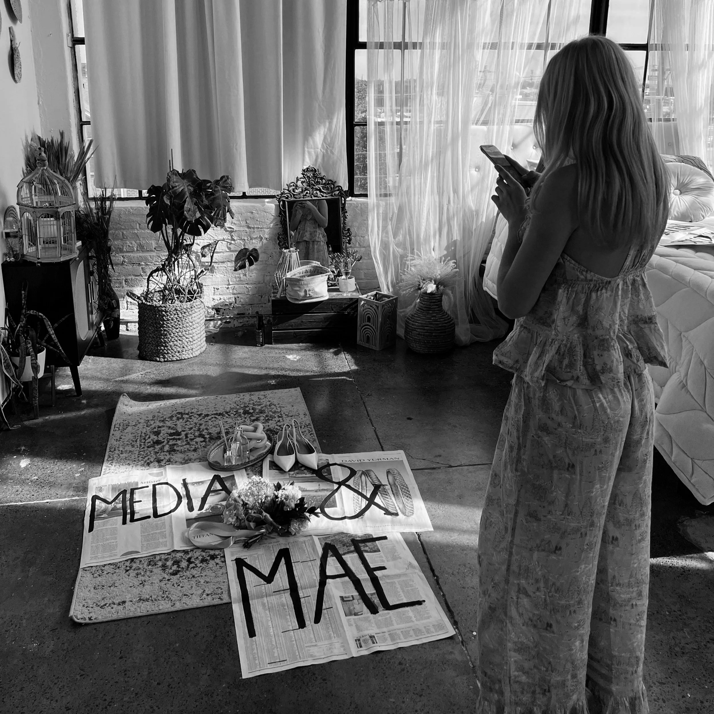 A woman standing on a concrete floor taking a photo with her phone, in a room with industrial-style brick walls, large windows, and sheer curtains. There are various decorative items, including a mirror, plants, and a birdcage, with a newspaper on the floor displaying the words 'MEDIA & MAC' and a pair of high heels and flowers on top.