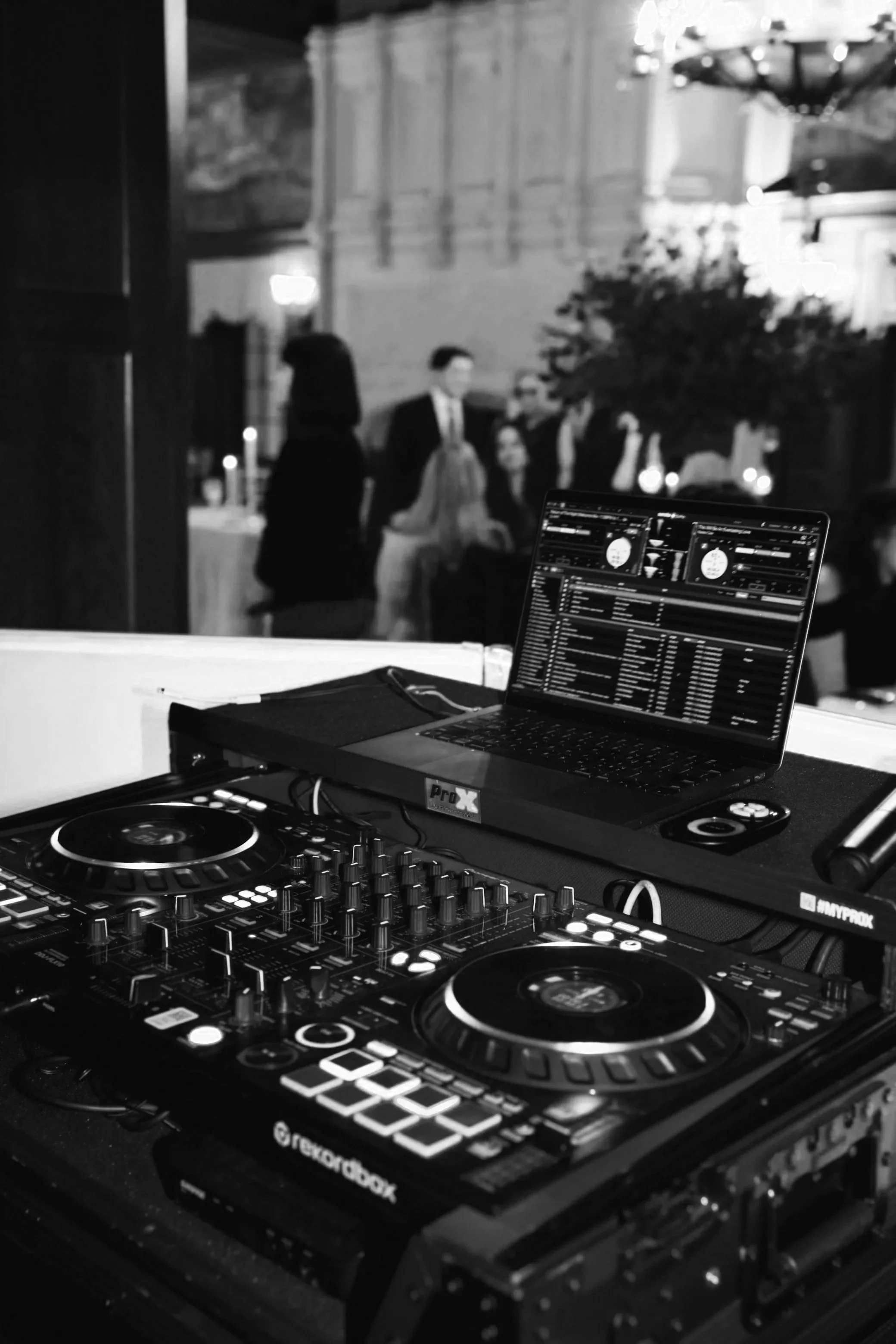 DJ setup with turntables, mixer, and a laptop at a party or event, with blurred guests in the background.