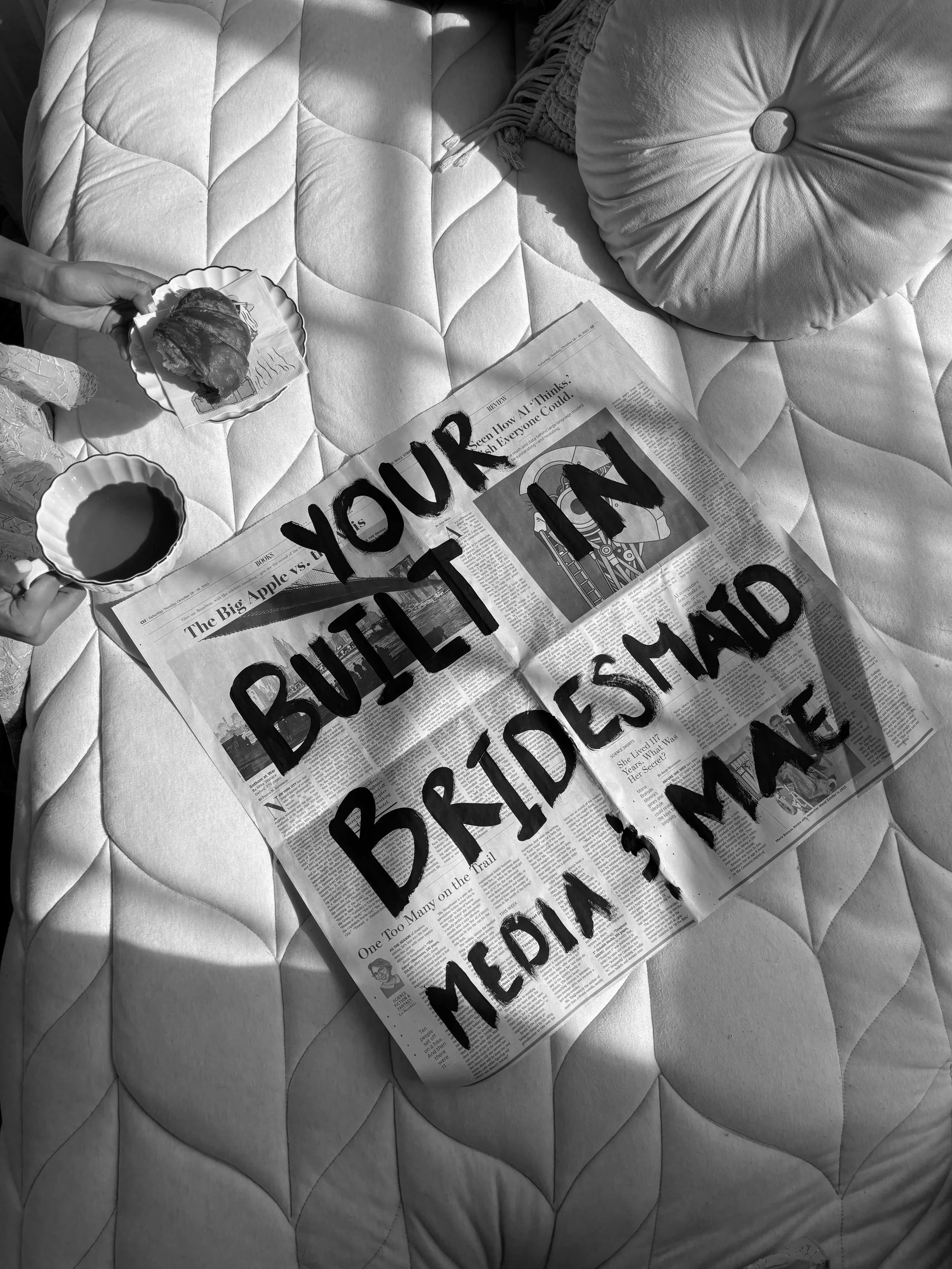 A bed with a quilted mattress, a round cushion, and a newspaper with a message written on it. The message says, "YOUR BUILDING BRIDESHAD MEDIA & MAE." There are two small bowls, one filled with a liquid and the other empty, and a person is holding a pastry and a cup near the newspaper.