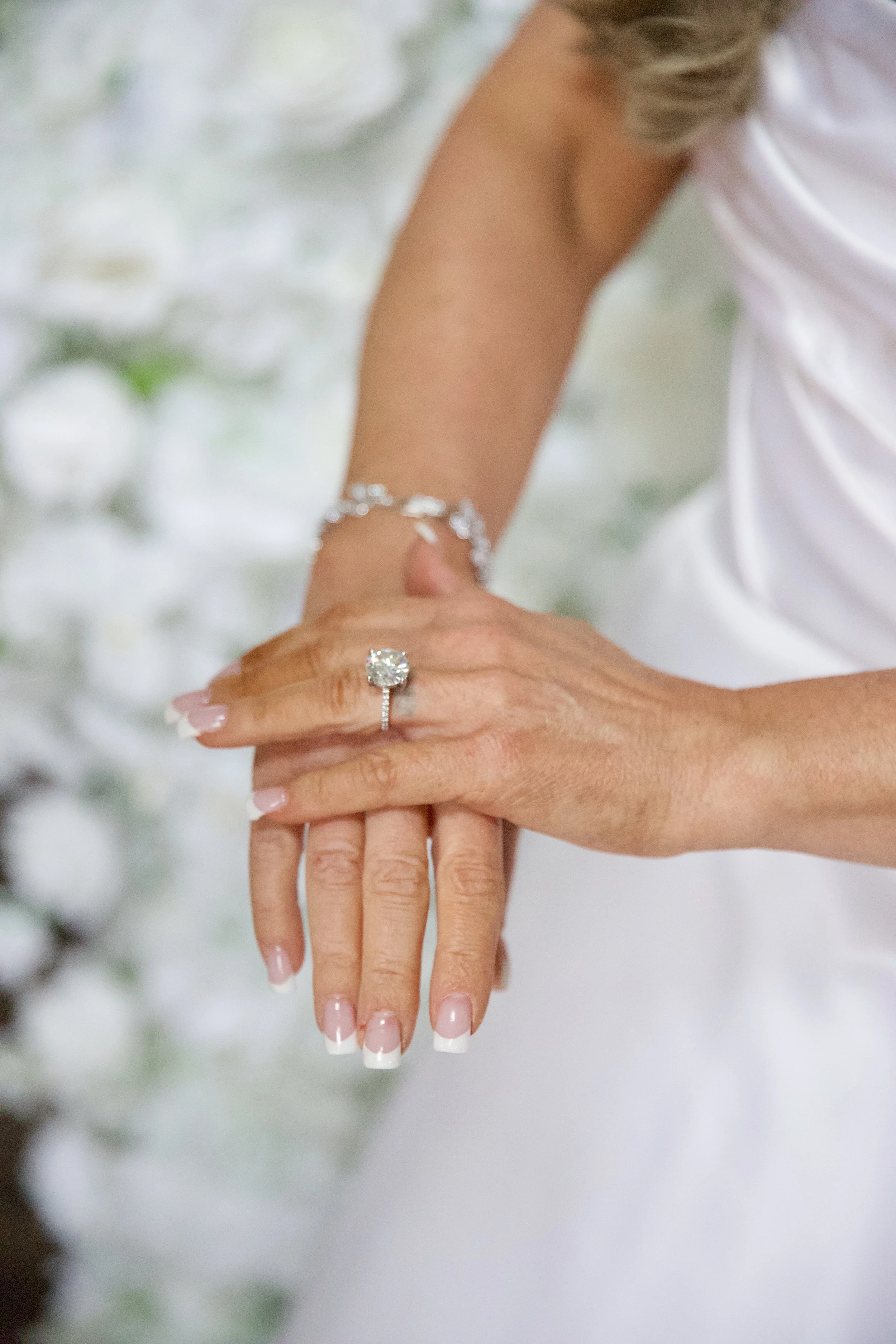 Close-up of a woman’s hands with an engagement ring and bracelet, with a white dress and floral background.