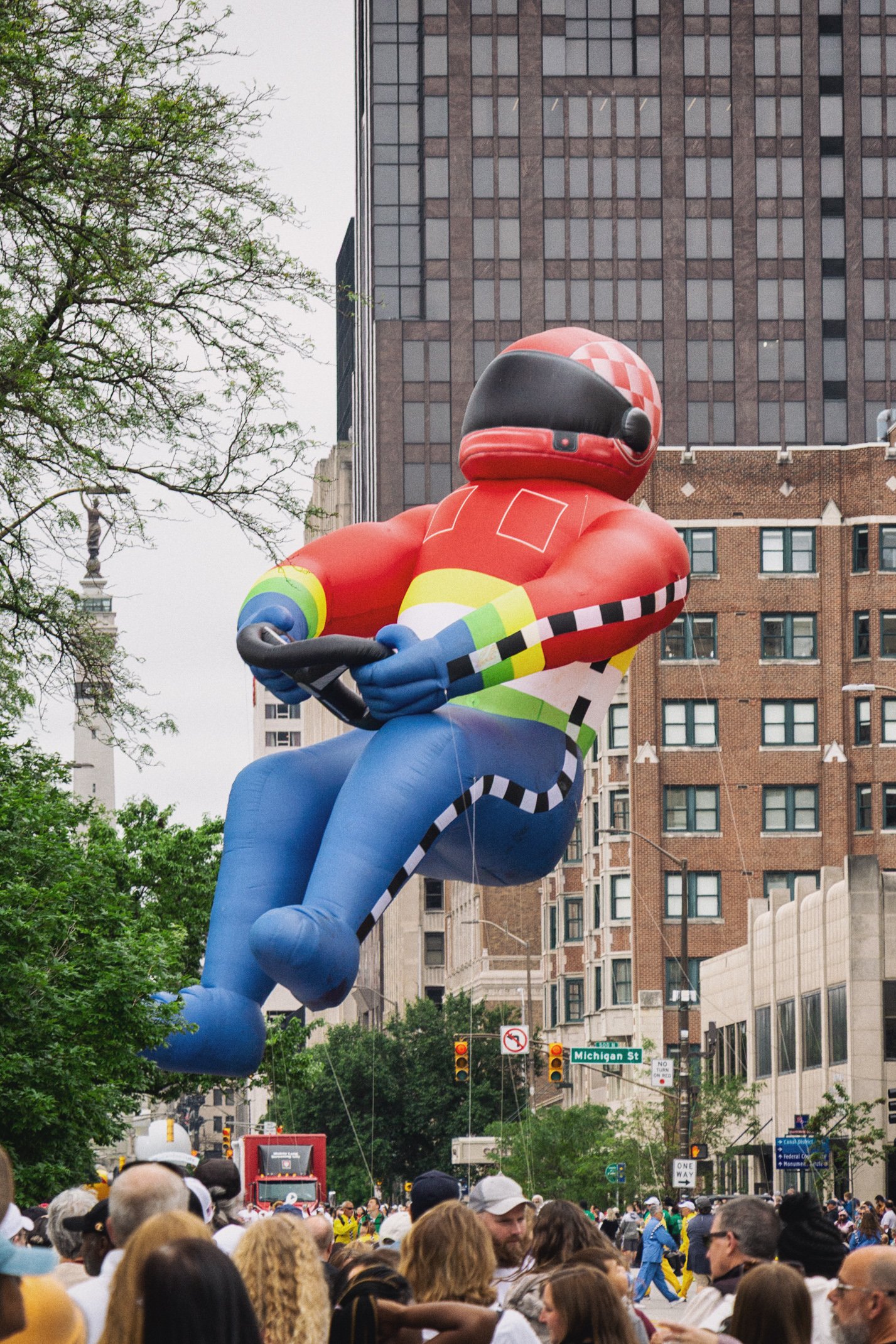Crowd of people gathered on a city street with tall buildings, traffic lights, and street signs, including Michigan Street, in the background. A large inflatable figure of a racing driver in red, blue, and yellow is floating above the crowd.