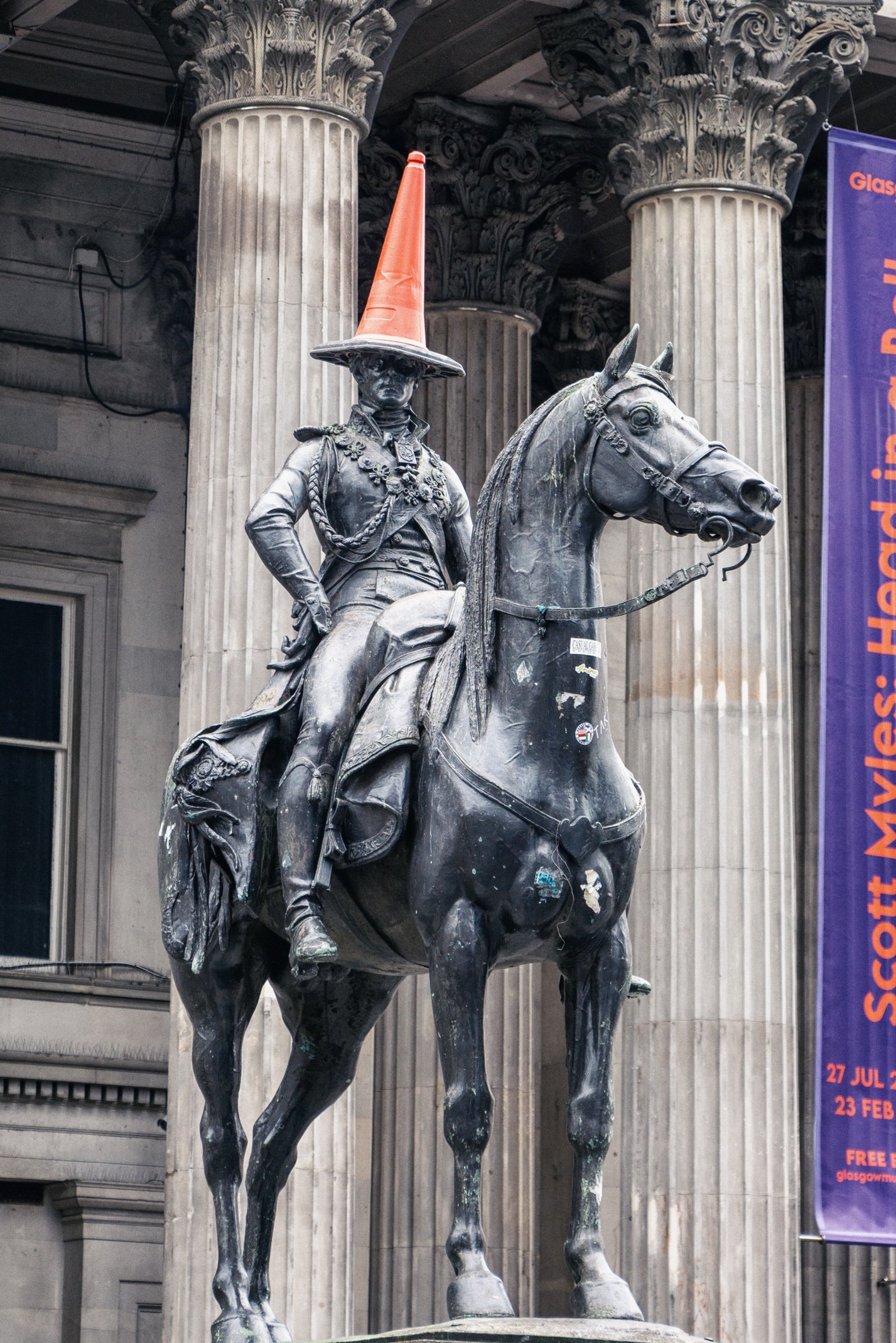 Statue of a military figure on a horse with an orange traffic cone on the head, situated in front of ornate columns.