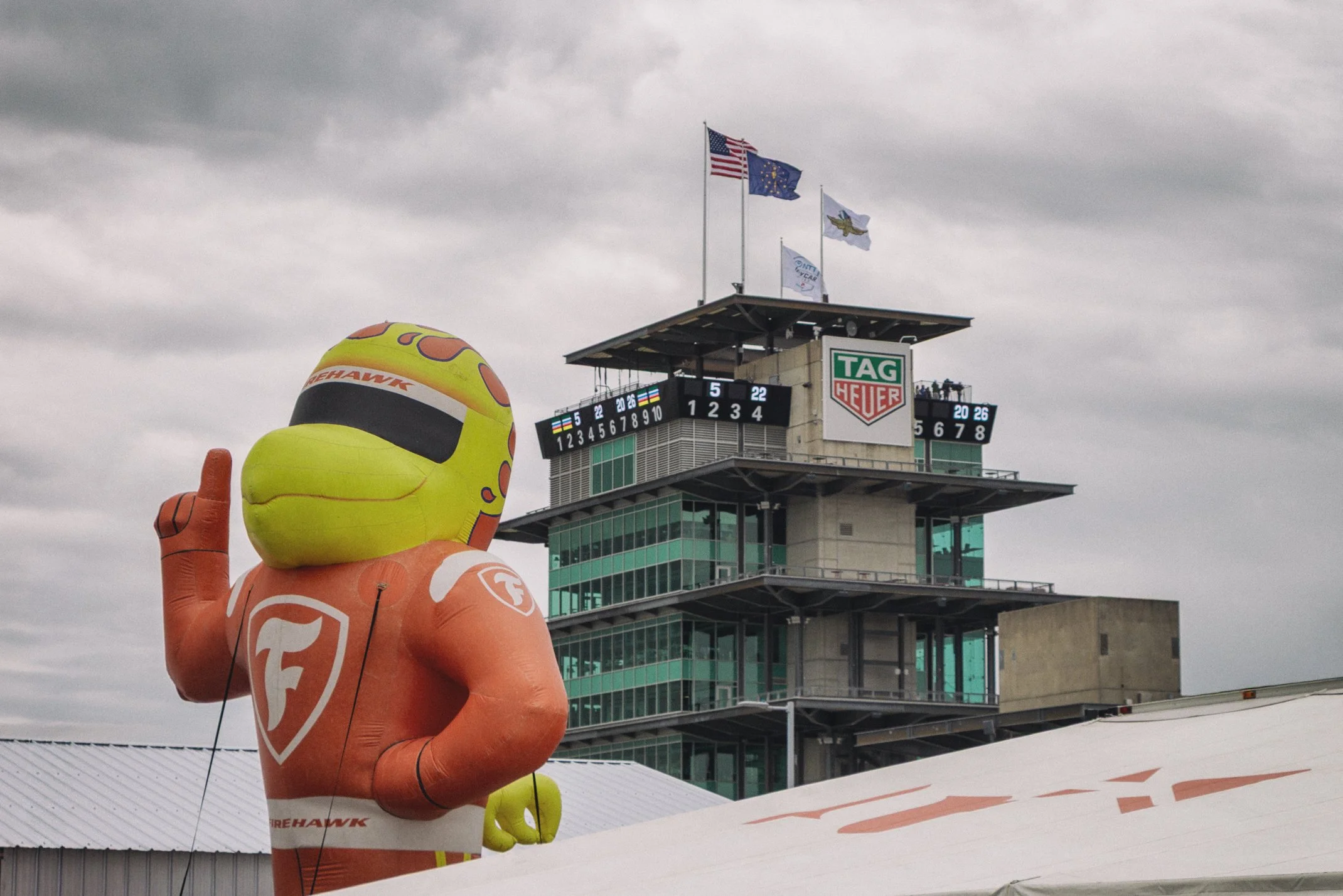Large inflatable mascot of an orange, with a yellow face and black eyes, wearing an orange racing suit with 'FIREHAWK' logo, at a race track.