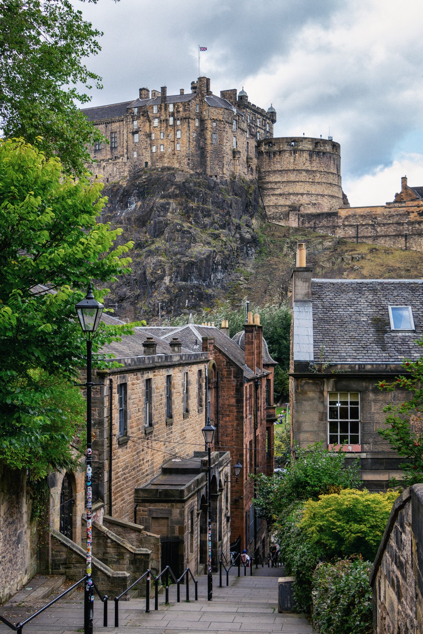 A street view of buildings leading up a hill with a castle on top, greenery on both sides, and cloudy sky overhead.