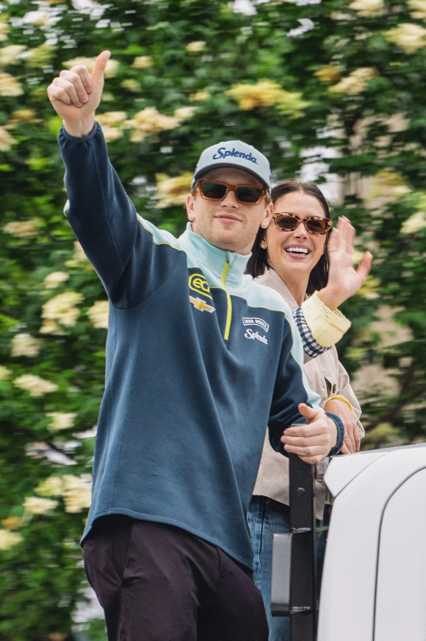 A man and a woman waving while riding in a golf cart with a blurred green leafy background.