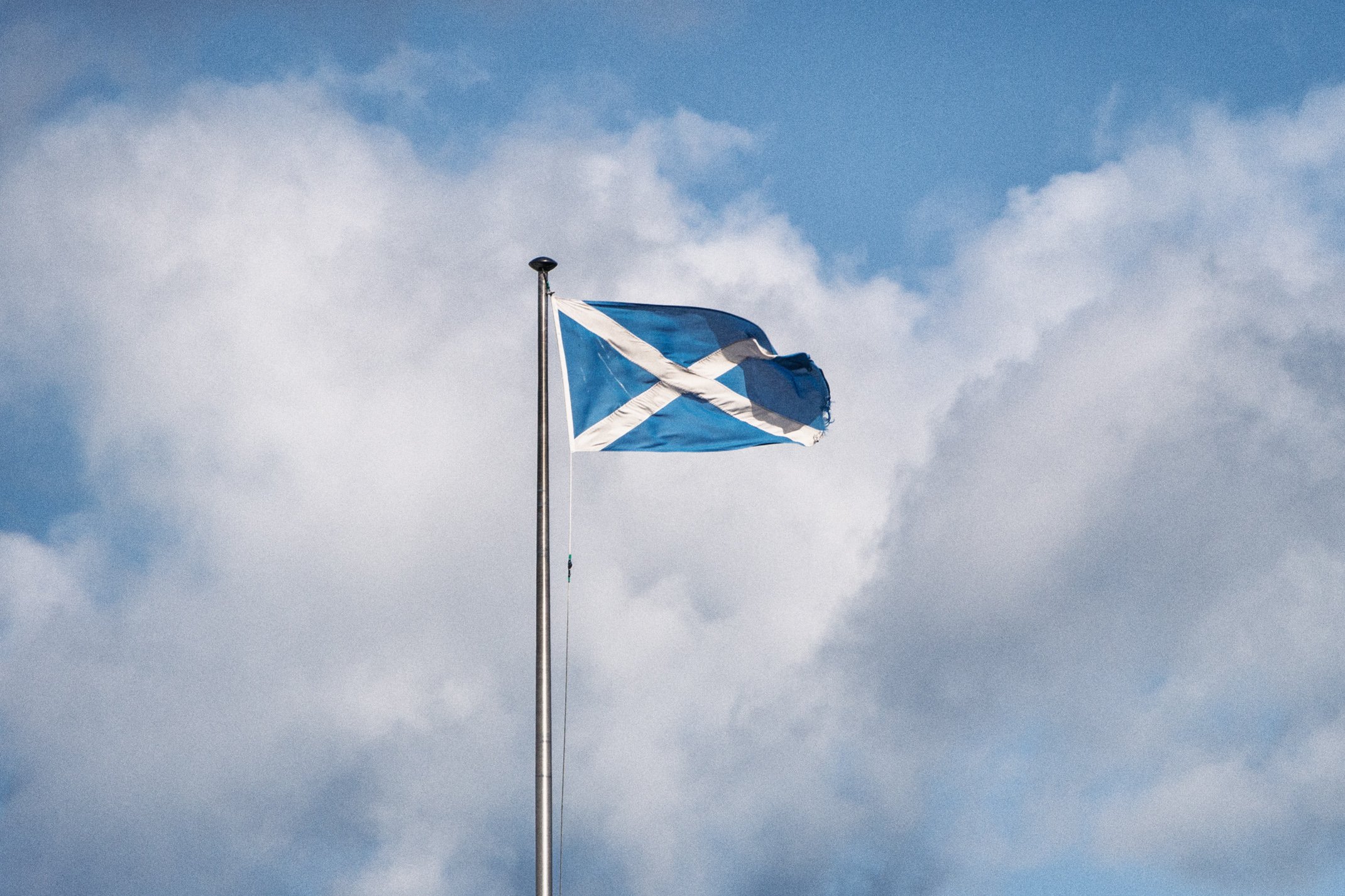 Scottish flag, known as the Saltire or Saint Andrew's Cross, waving on a flagpole against a sky with clouds.