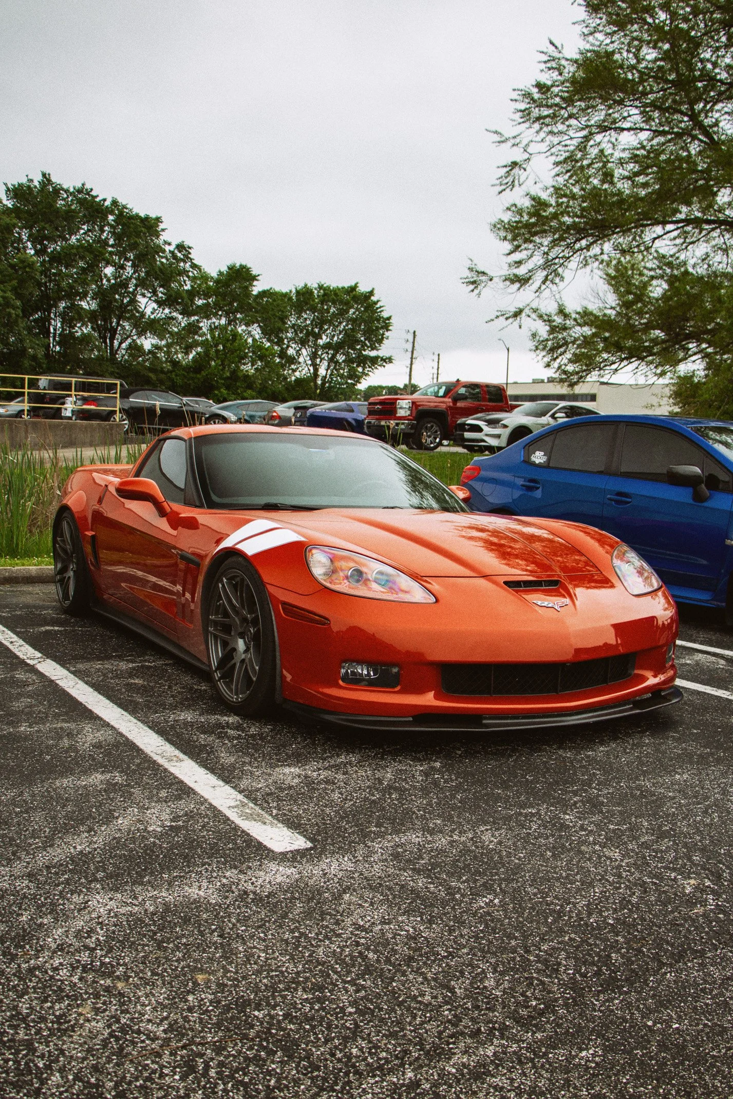 Orange sports car parked in a parking lot, with a blue car parked beside it and other vehicles in the background. Trees and overcast sky are visible.