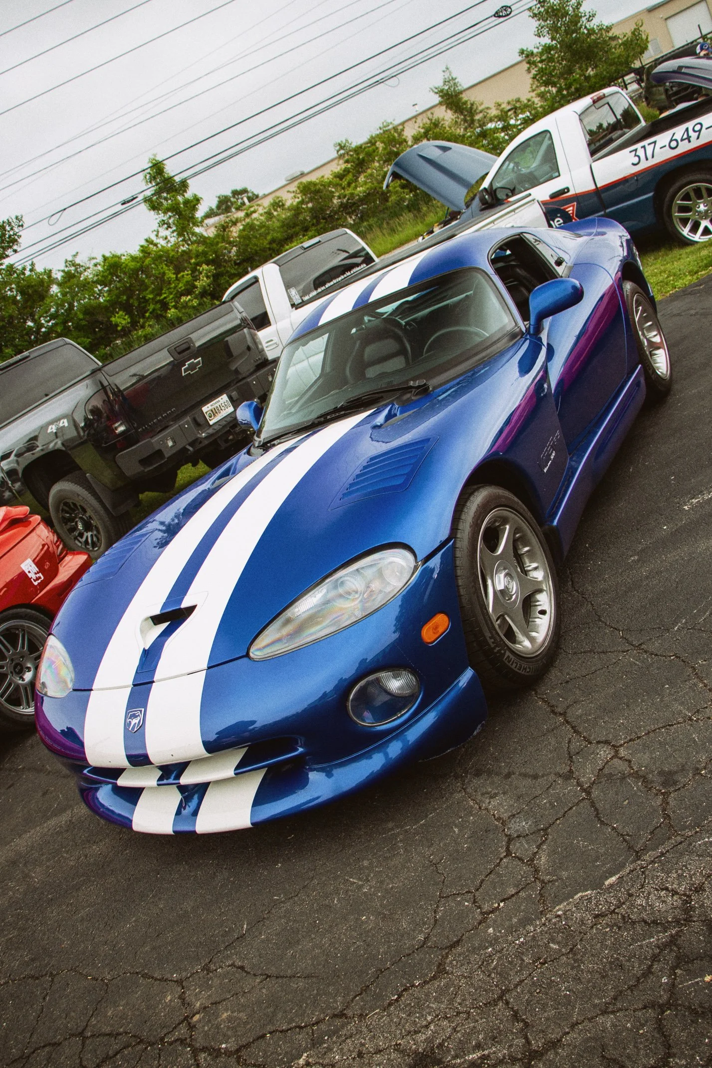 A blue sports car with white racing stripes, parked on a cracked asphalt lot. There is a white pickup truck with its hood open and a black pickup truck nearby, along with other vehicles. Trees and power lines are visible in the background.