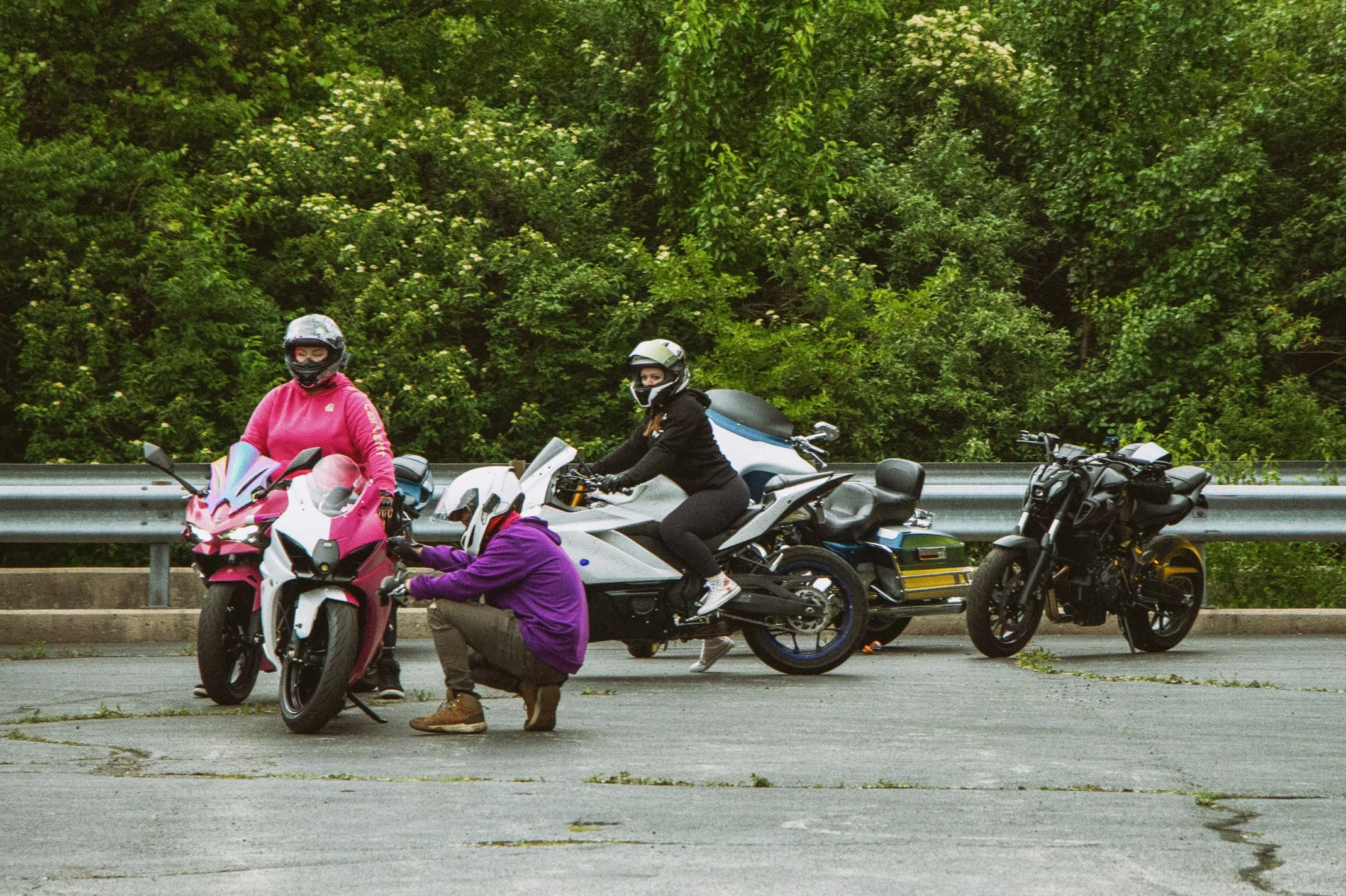 Four motorcyclists, two women and two men, wearing helmets and protective gear, stopped on the side of a road with a green, leafy background, and a person crouching near a motorcycle.