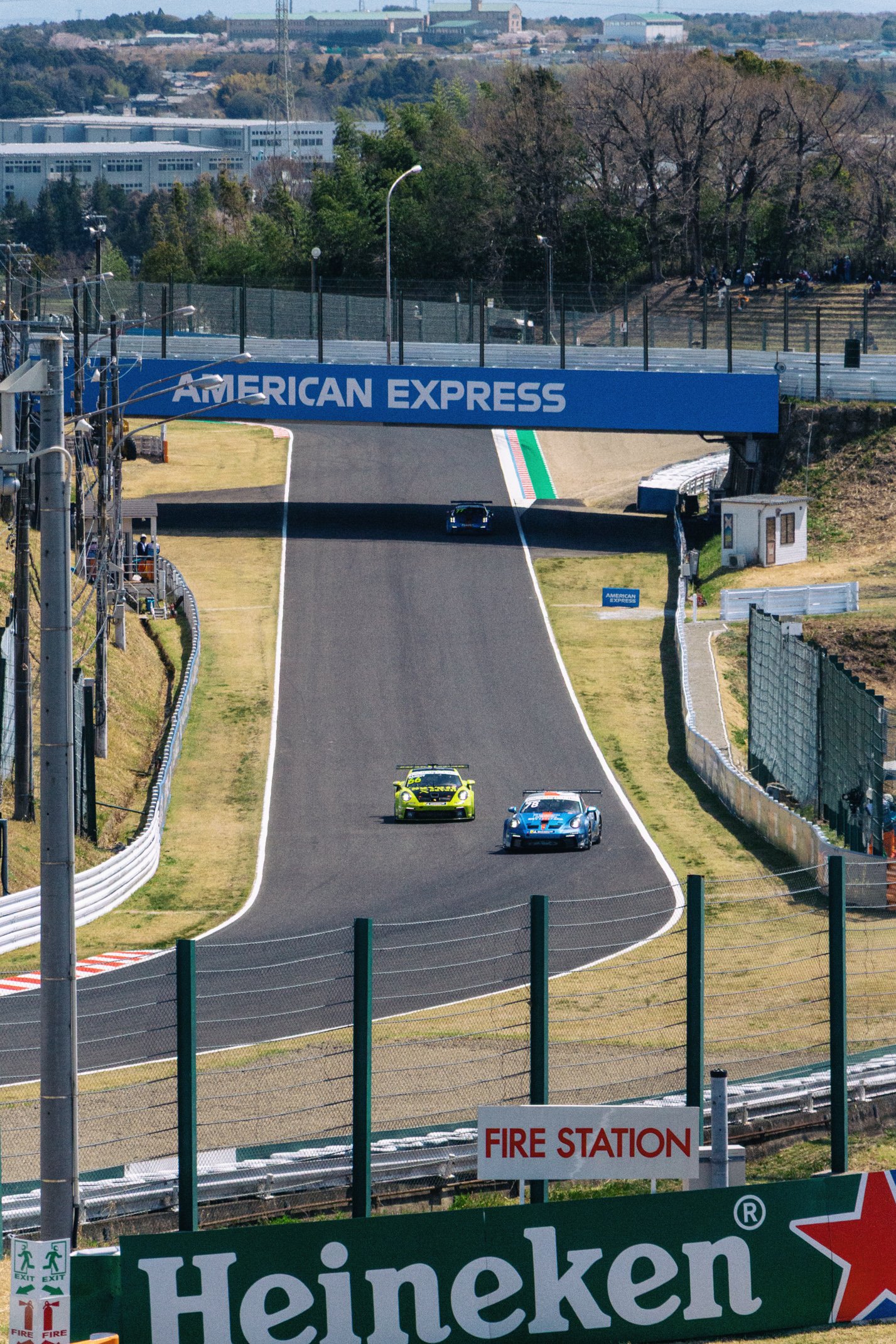 Race cars driving on a track under a large American Express sign overpass, with a fire station sign and a Heineken advertisement in the foreground.