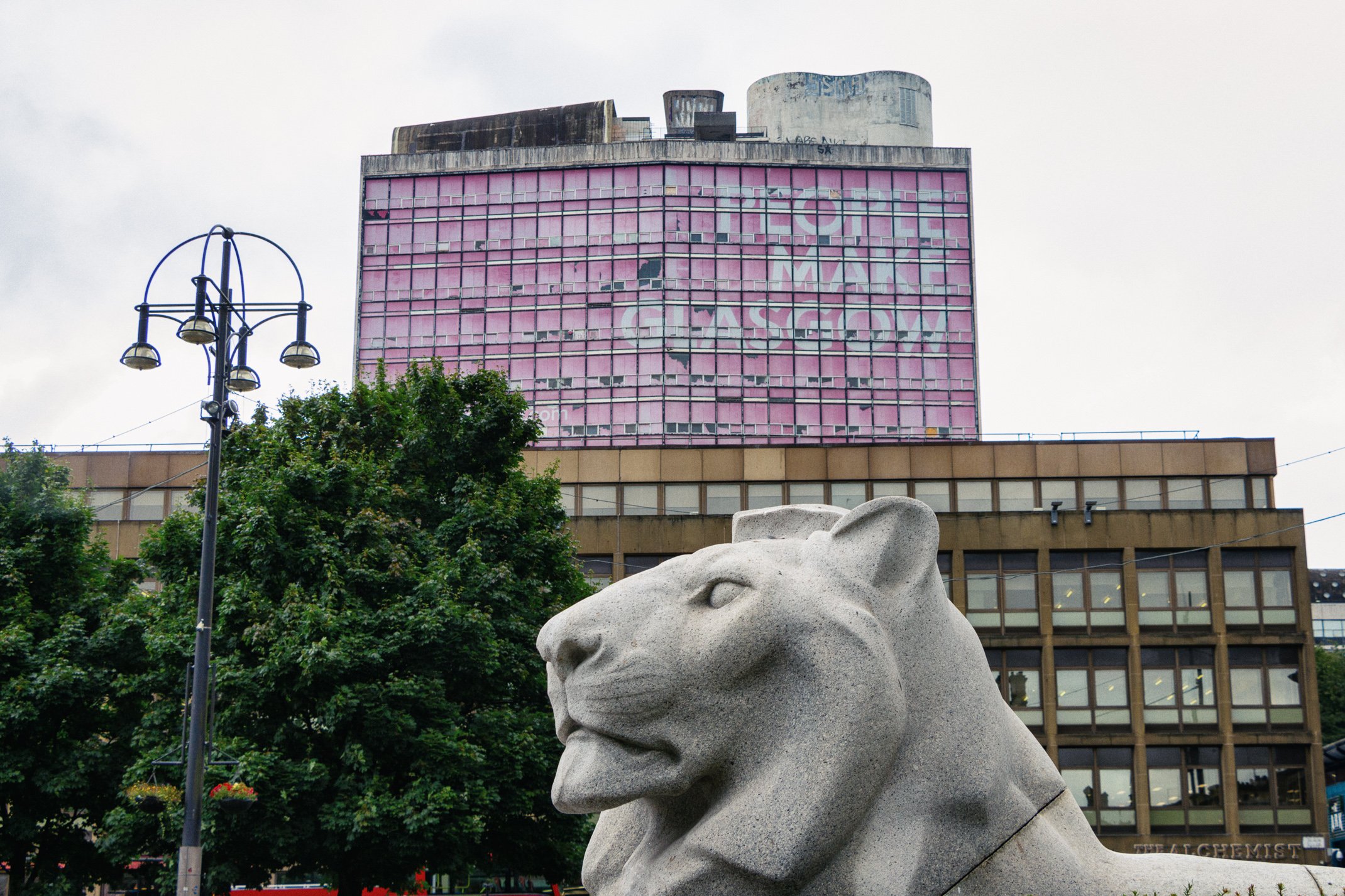 Statue of a lion in front of a multi-story building with pink windows and a large painted advertisement that reads "TOP MACK GASSOW". There is a tree with green leaves and a street lamp to the left.