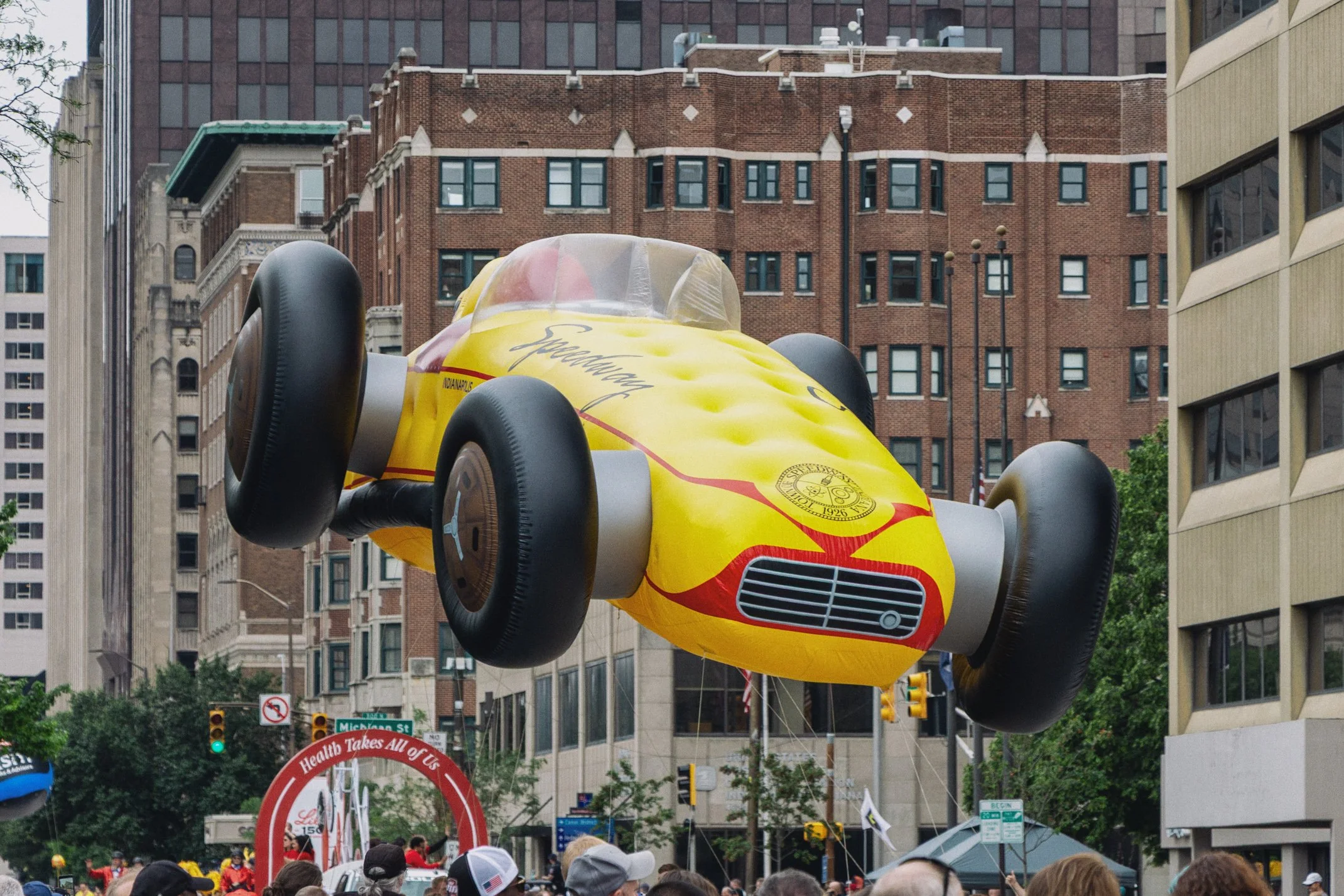 Situation room with a large yellow IndyCar race car floating above a crowd of people on city street, with high-rise buildings in background.